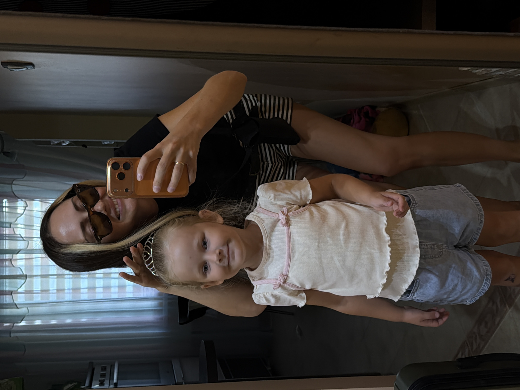 The image shows a woman and a young girl standing in front of a mirror, taking a selfie. The woman is wearing sunglasses, a black t-shirt, and striped shorts. She has blonde streaks in her dark hair. The young girl is wearing a white top with pink trim and denim shorts. She has a tiara on her head. They are in what appears to be a home environment, possibly a hallway or room with a mirror on the wall. The background includes curtains, a table, and a door. There is no visible indication of the city of Kamyshin, Russia, or any other specific location.