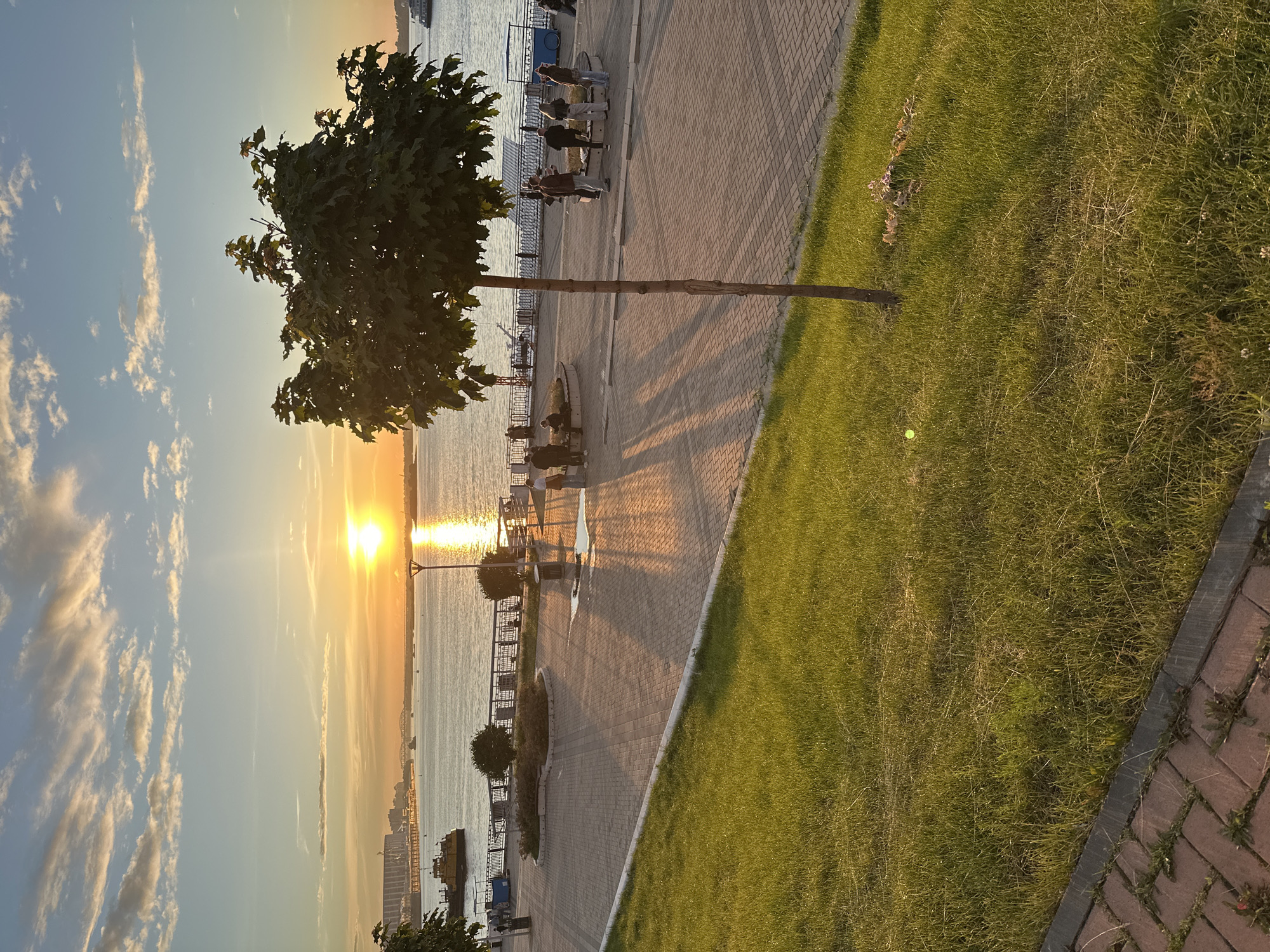 An outdoor, eye-level, vertical shot captures a sunset over a wide river. The sun, a bright white orb, is positioned in the upper left quadrant of the sky, casting a golden reflection on the water and creating long, sharp shadows across the paved walkway in the foreground. The sky is a gradient of blues and oranges, with wispy clouds.
On the left side of the image, a strip of vibrant green grass slopes down from the foreground, bordered by a brick pathway. Beyond the grass, a paved promenade runs along the riverbank, lined with a dark metal railing. A few small, decorative bushes are planted behind the railing. Further back on the promenade, several people are standing and walking, some gathered in small groups. One individual, wearing a white top and light-colored pants, stands near the railing. Another group of people are conversing near a bench. A lone, slender tree with dark green leaves stands in the mid-ground, its shadow stretching across the walkway. In the distance, on the river, a yellow tugboat is visible, along with several other smaller boats. Across the river, a long building or structure with a light-colored facade and a dark roof is discernible.
On the right side of the image, the promenade continues, with more people strolling. One person in a dark outfit stands facing the river. The scene is bathed in the warm light of the setting sun, creating a serene and tranquil atmosphere.