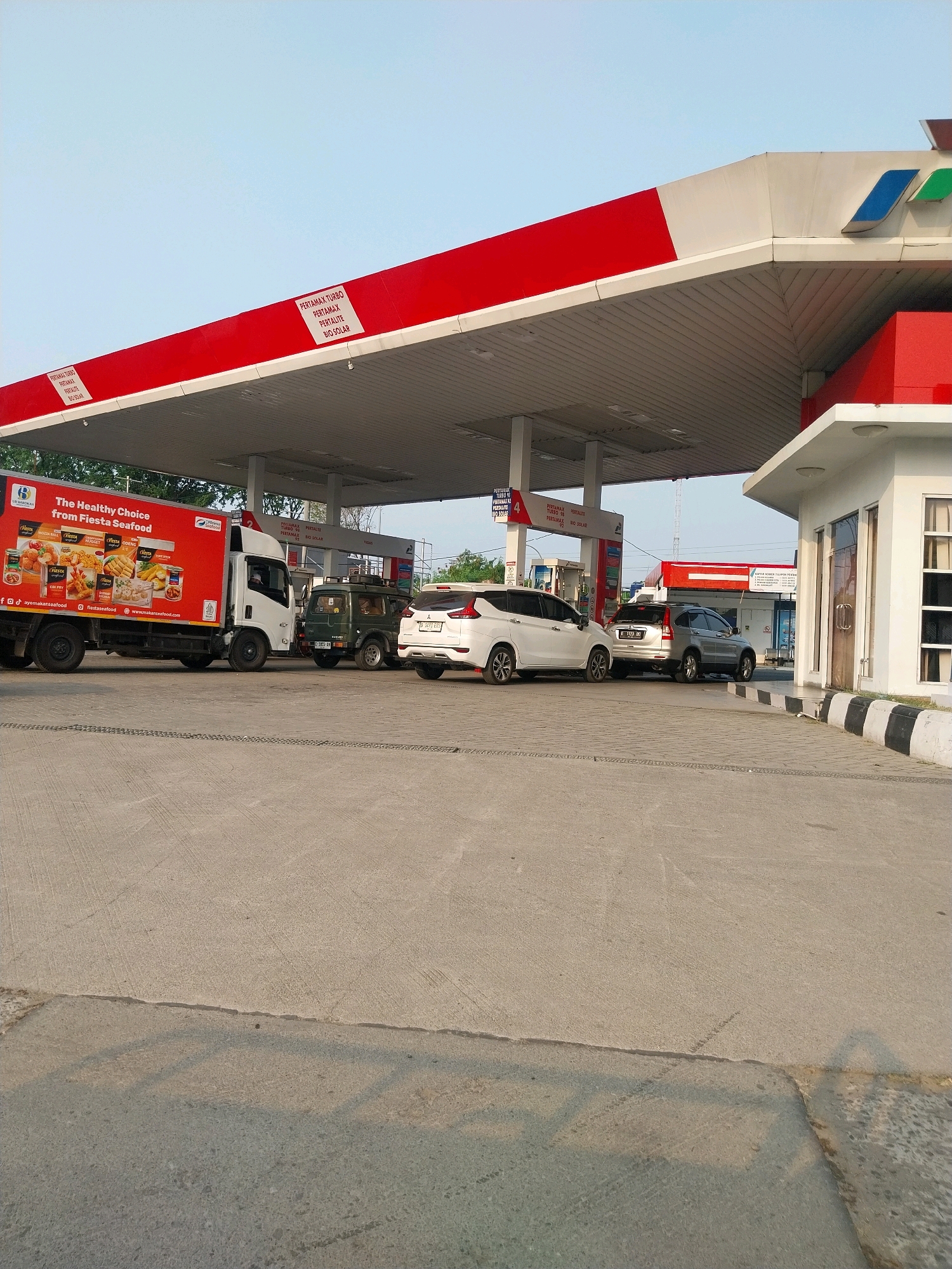 The image depicts a gas station in Cirebon, Indonesia. The station features a red and white canopy with the Pertamina logo, a prominent fuel company in Indonesia. Several vehicles are present, including a white delivery truck with "The Healthy Choice from Fiesta Seafood" branding, a green SUV, and two white cars. The scene appears to be an active fueling operation. The environment is an open-air paved area with a clear sky overhead, suggesting daytime. The gas station's buildings and pumps are visible, as well as signage indicating fuel types like Pertamax Turbo, Pertamax, Pertalite, and Bio Solar. The overall impression is a typical commercial scene at a gas station.