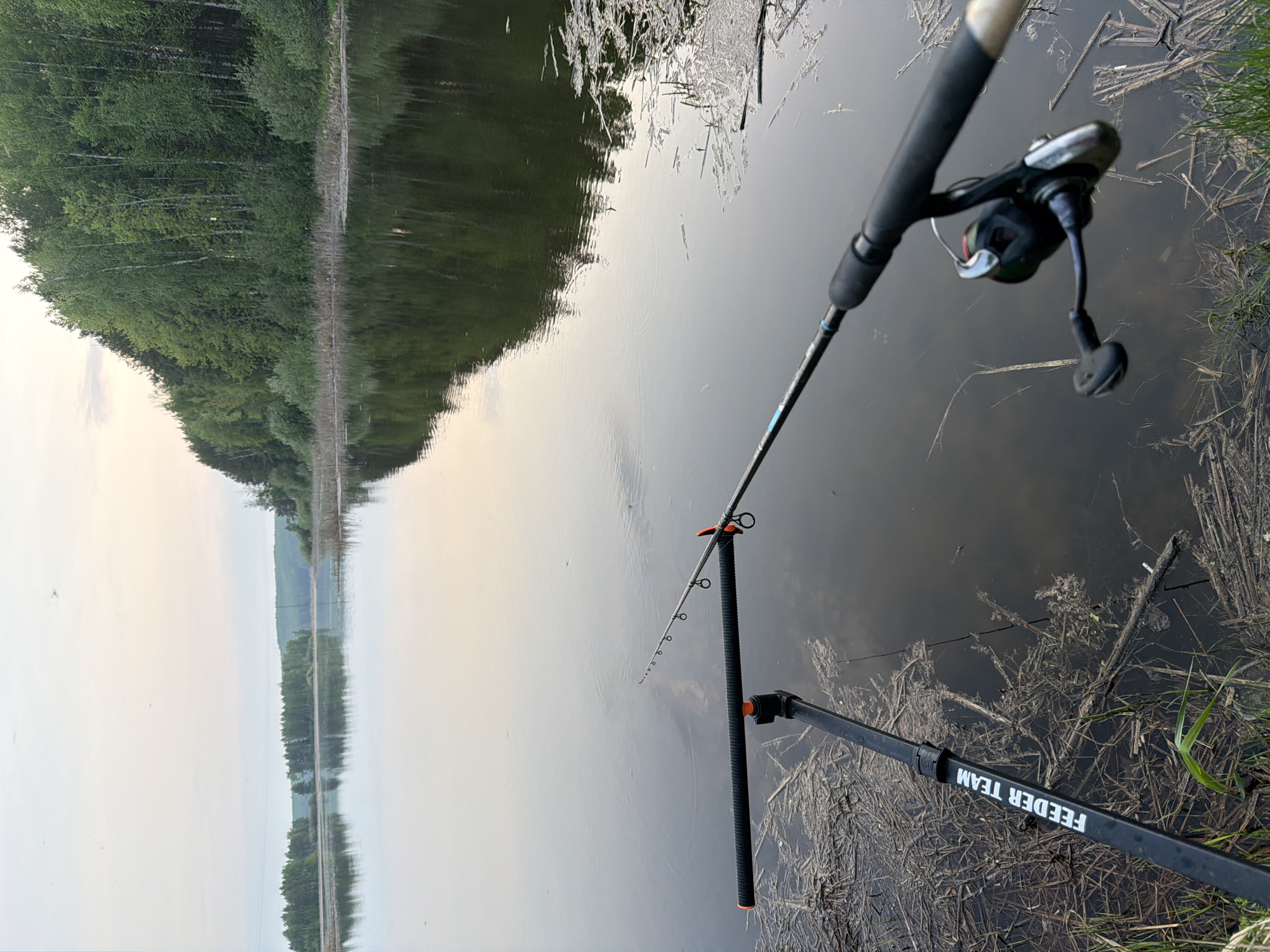 The image depicts a fishing scene at a water body, likely a lake or a calm river, in Chaykovsky, Russia. A fishing rod is set up on a rod rest, with the rod extending into the water. The reel is attached to the rod. Debris, including dead leaves and small branches, float on the surface of the water and are also present on the bank. A line of trees is visible on the opposite bank, with their reflection visible in the water. The sky is overcast. The words "FEEDER TEAM" are printed on the rod rest.