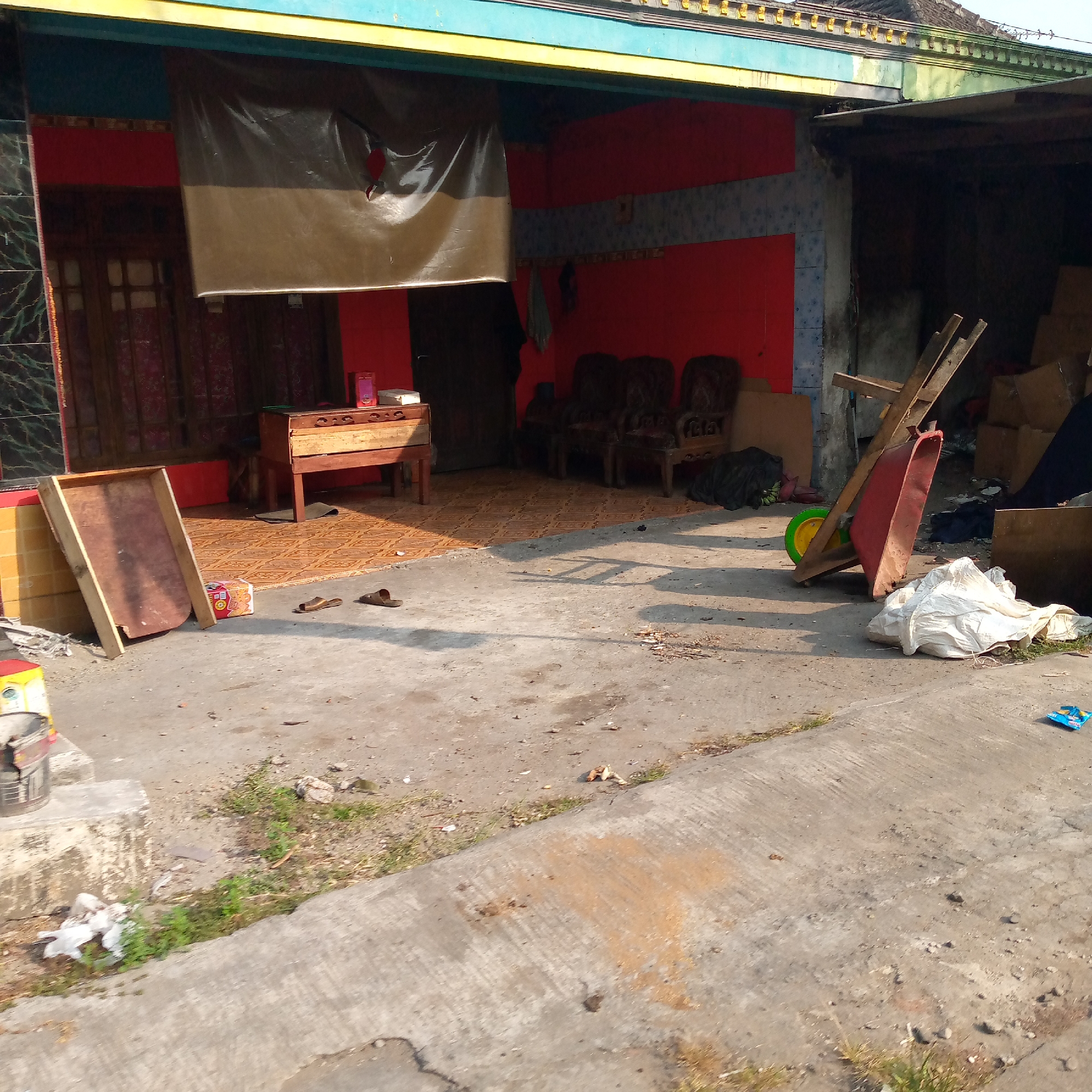 The image depicts an outdoor scene in Tambakagung, Indonesia. In the foreground, a concrete ground is visible with scattered debris, including a blue packet, a piece of white plastic, and some vegetation. A rough concrete barrier with grass growing alongside it extends into the midground. Further back, a wooden structure leans against a tiled wall. To the right, a red wheelbarrow with a yellow wheel is positioned near a pile of white plastic sheeting. Behind the wheelbarrow, there are stacks of cardboard boxes, some covered by dark material. The background shows a building with a red wall, a dark tarp hanging over an opening, and a wooden table with items on top. Three ornate wooden chairs are visible in front of the building. The overall scene suggests an outdoor storage or work area adjacent to a dwelling.