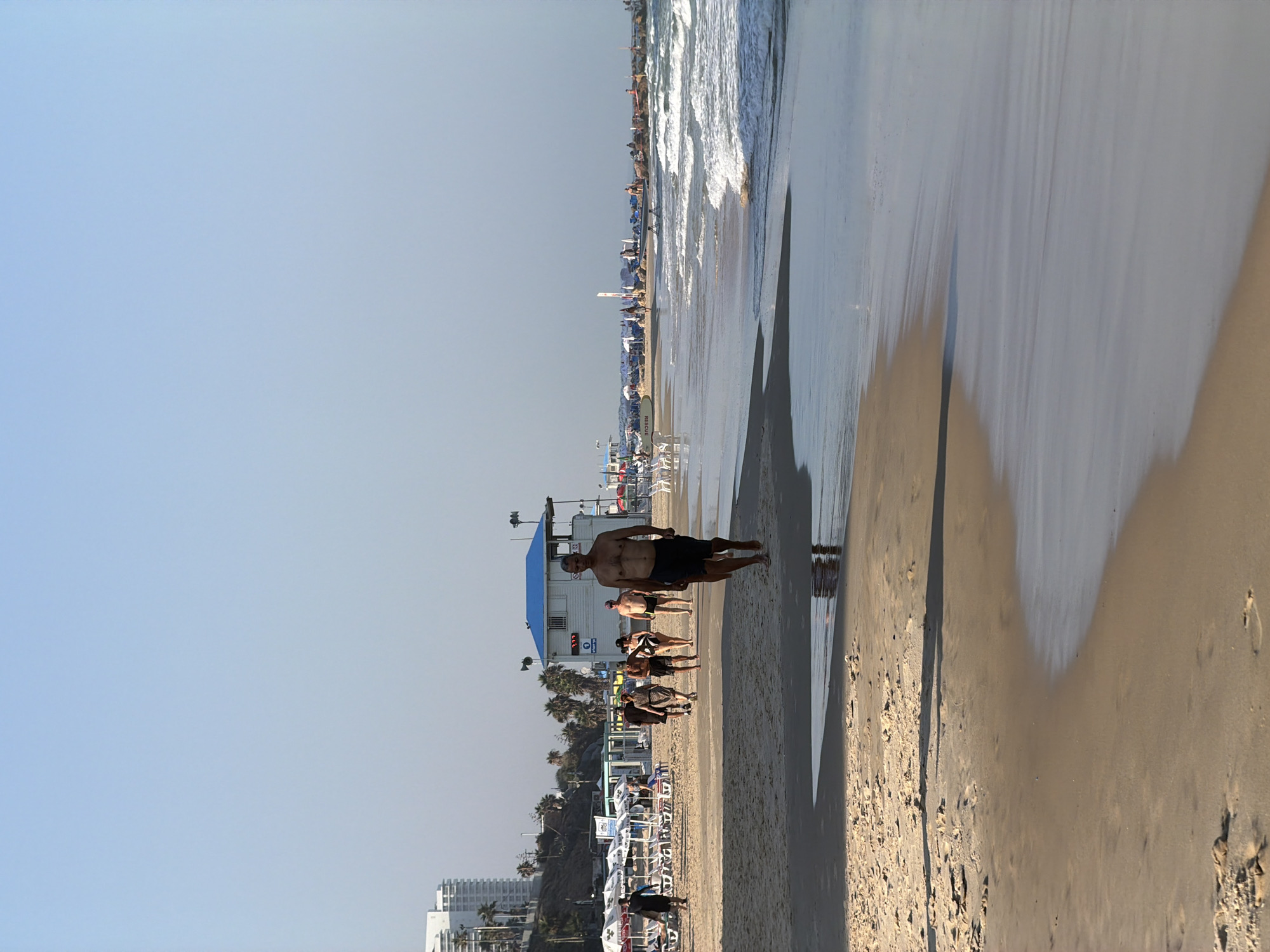 This is a photograph of a beach in Bat Yam, Israel. In the foreground, a man walks along the wet sand near the water's edge, with his shadow cast on the beach. Several other people are walking on the sand further back, some in groups. Behind them, there is a lifeguard station with a blue roof, beach chairs, and umbrellas. In the distance, buildings and palm trees are visible along the coastline, and the sea stretches out to the horizon. The sky is clear and pale blue.