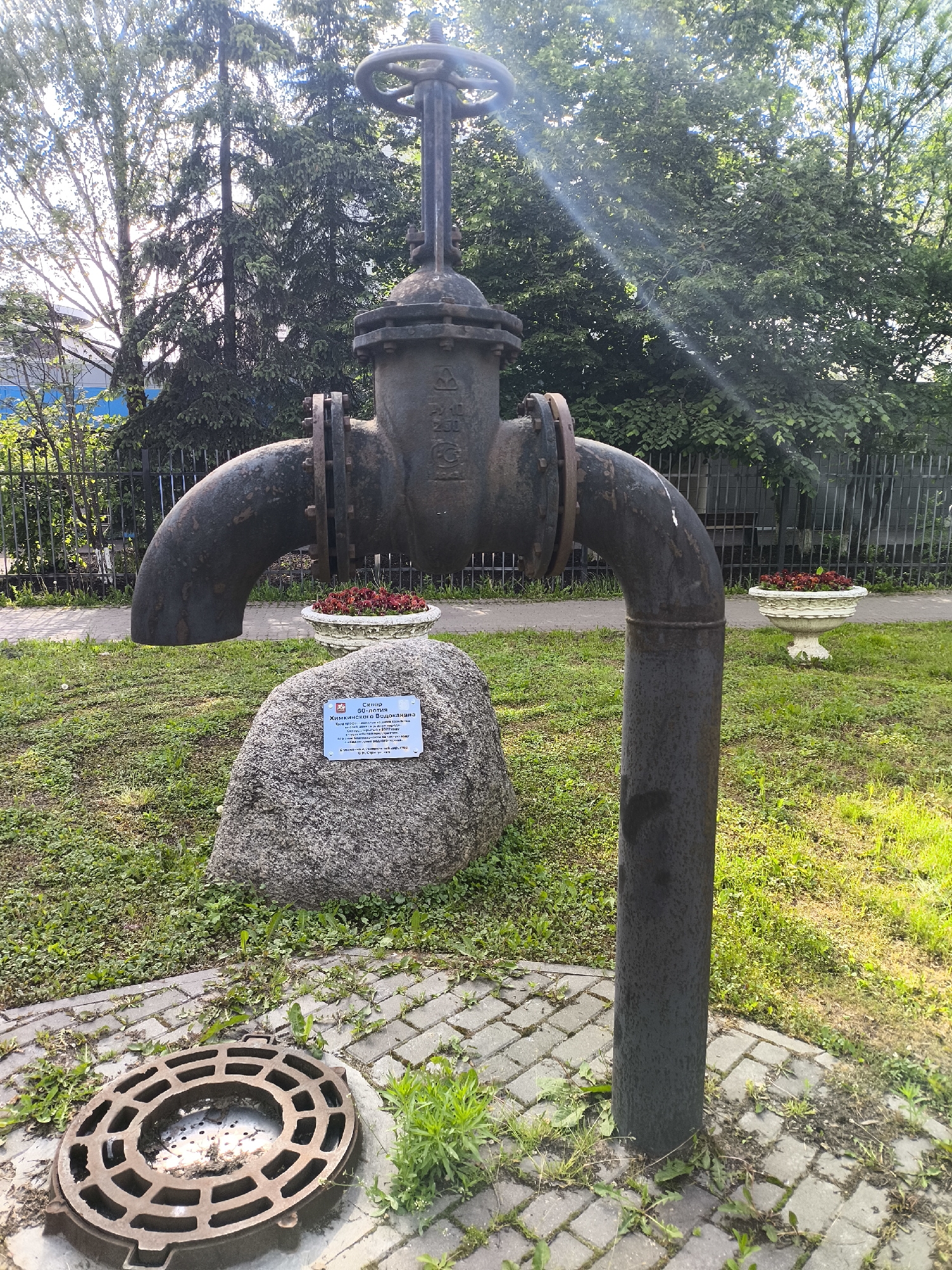 A metal pipe sculpture with a wheel valve sits atop a cobblestone pavement in a grassy area near trees and a black metal fence. A large grey stone with a plaque is positioned in front of the sculpture. A round metal grate lies on the pavement in the foreground. Two flowerpots with red flowers are visible near the sculpture. The location is Unknown, Unknown.
