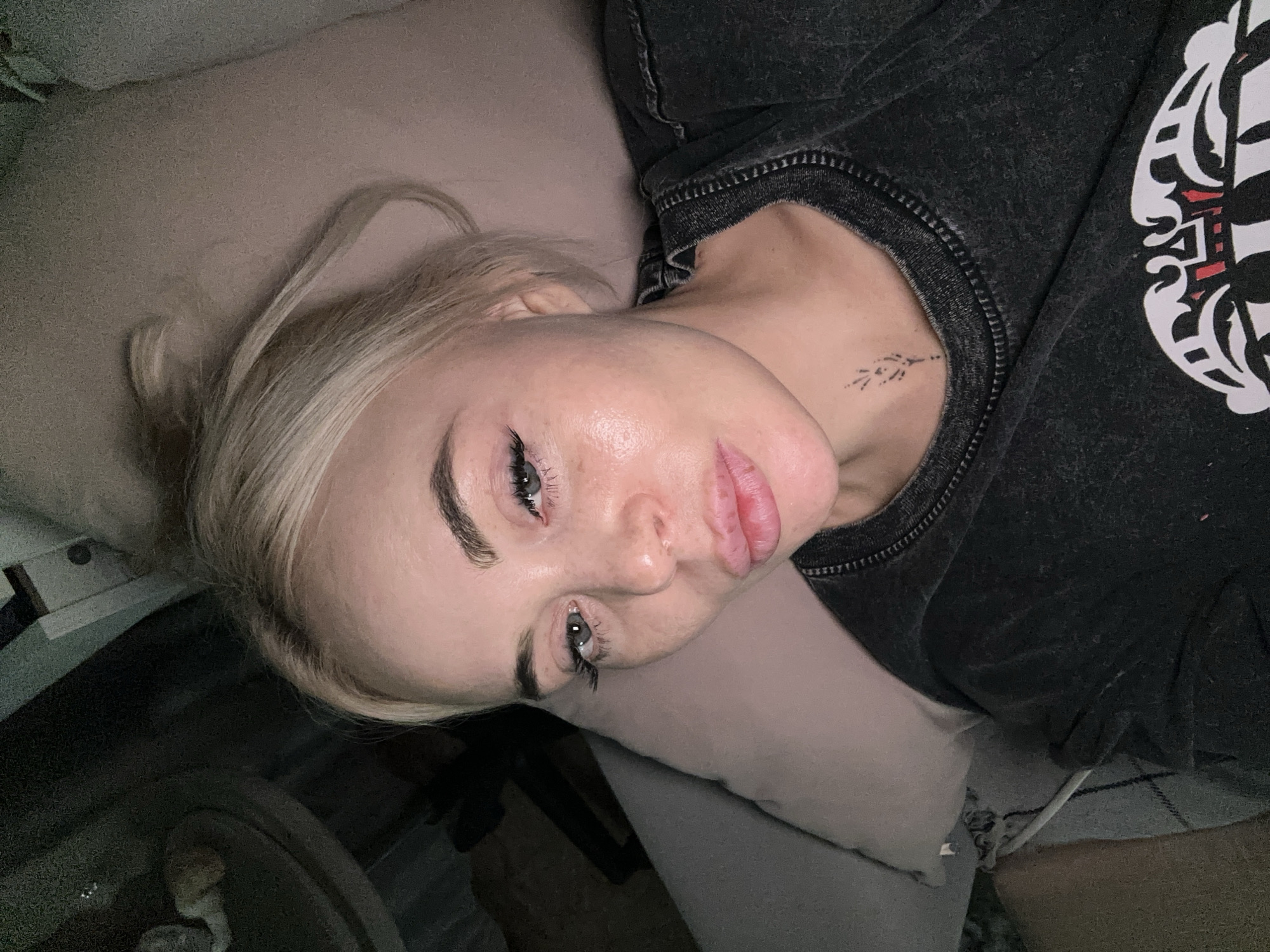 A young woman with blonde hair lies on a light-colored pillow. She is wearing a dark-colored shirt with a design on the upper right side. There is a small tattoo on her upper chest/neck area. The environment includes a blurred background with a possible table and other objects. The location is Высокий, Russia.