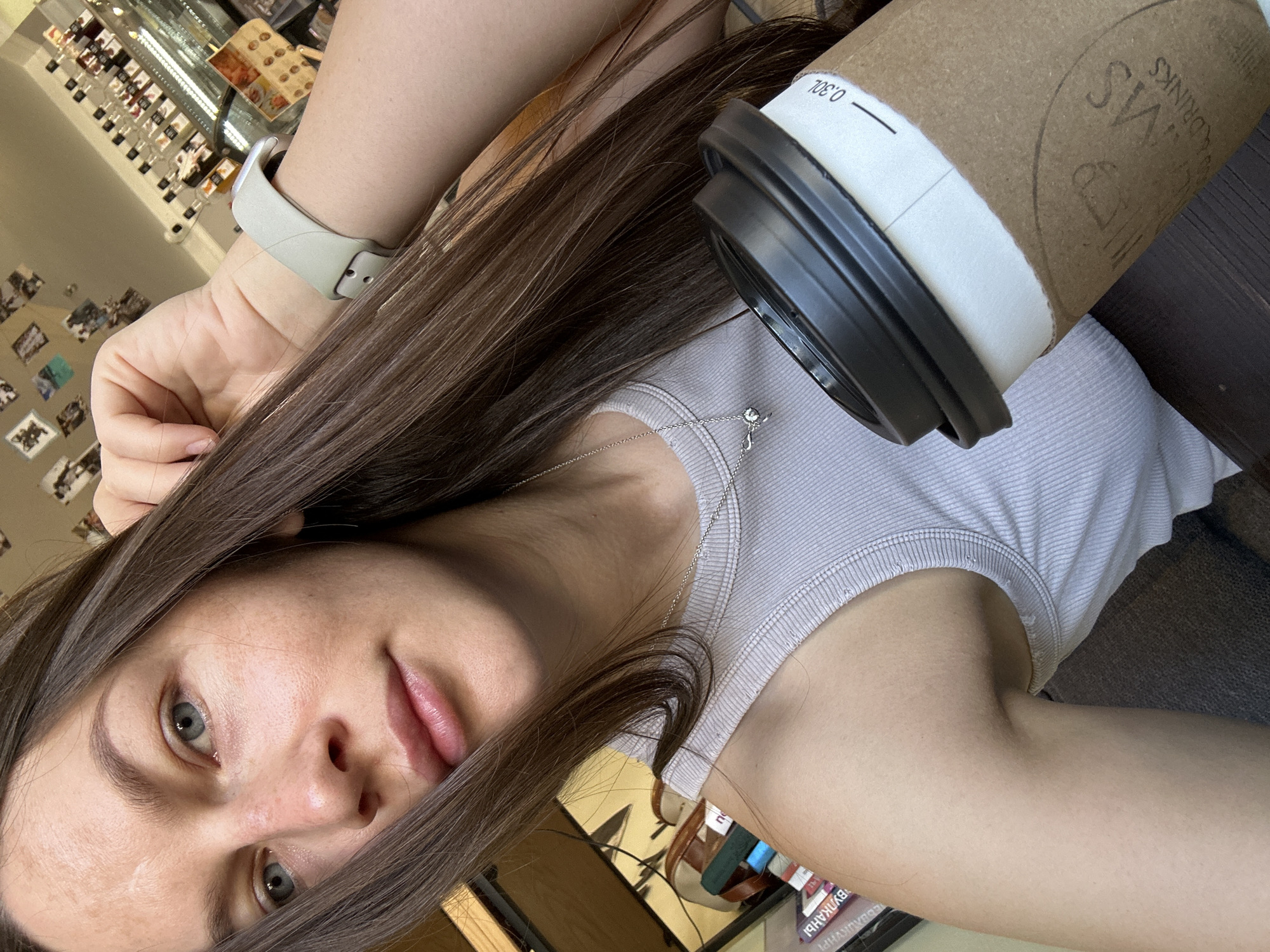 A woman with long, straight, brown hair and fair skin is holding a cup of coffee. She is wearing a light gray tank top and a light-colored smartwatch. She also has a silver necklace with a pendant. The coffee cup is brown and white with a black lid. The cup appears to be from a shop named "VS Coffee & Drinks" and the woman is likely located in Budva, Montenegro. The background shows a cafe interior, including shelves of items and photographs on a wall. The image is taken from a high angle, giving a view of her face, upper body, and arm.