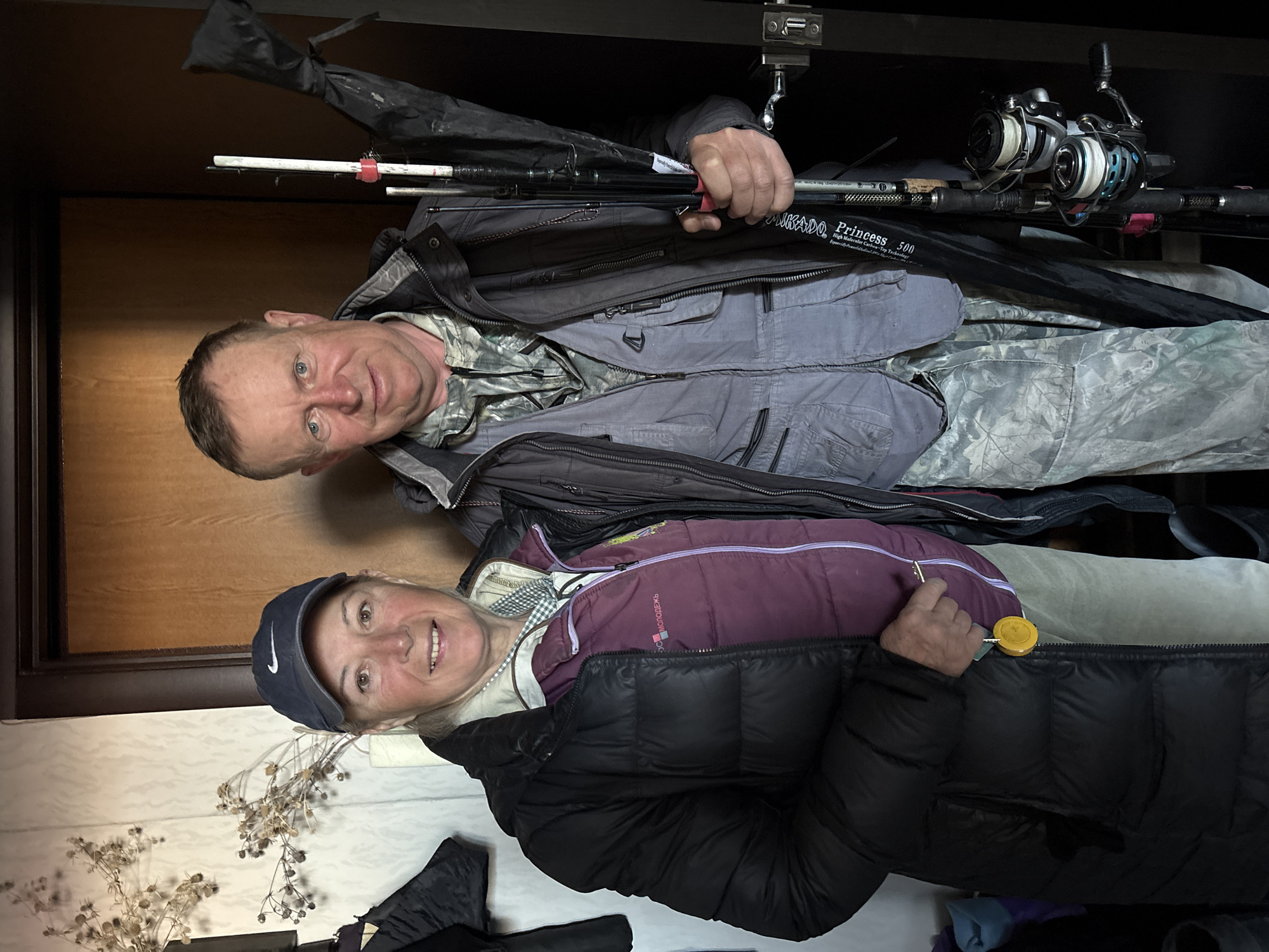 Two people pose indoors in Ulyanovsk, Russia, apparently preparing for or returning from fishing. A man in a grey jacket with a camouflage-patterned shirt underneath holds several fishing rods with reels. A woman in a black puffer jacket and blue baseball cap stands beside him, holding a small yellow object in her hand. The background suggests they are in a closet or storage area, with additional jackets and fishing equipment visible. Dried flower stalks are on the left side of the image.