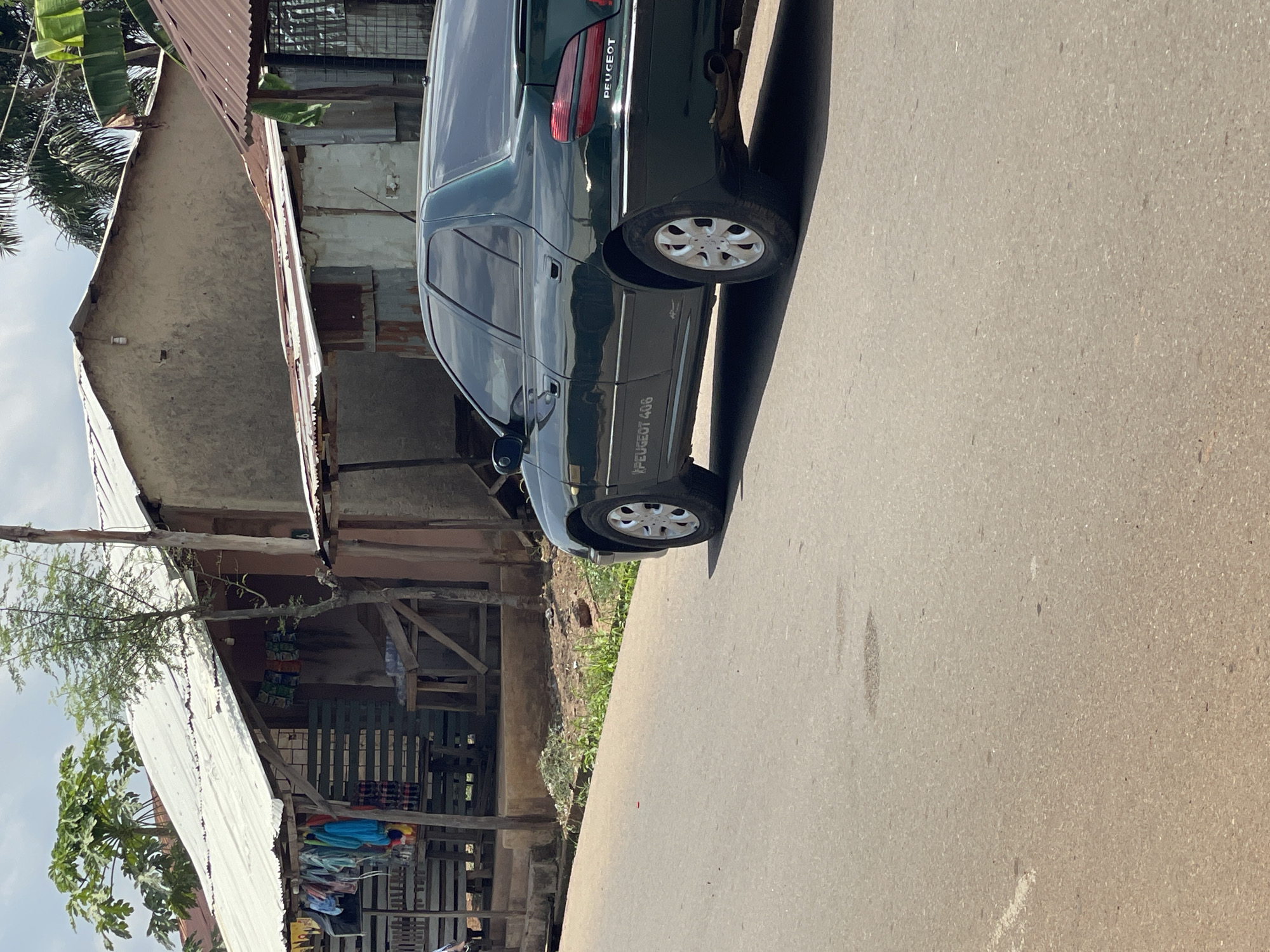 The image shows a dark green Peugeot 406 parked on a paved road in what appears to be a residential area in Akwanga, Nigeria. The car is positioned close to the side of the road, adjacent to a weathered building made of mixed materials including metal sheets and timber. The building appears to be a shop. The road is mostly devoid of other vehicles or pedestrians, emphasizing the stillness of the scene. The background shows hints of foliage and other buildings, giving context to the Akwanga setting.