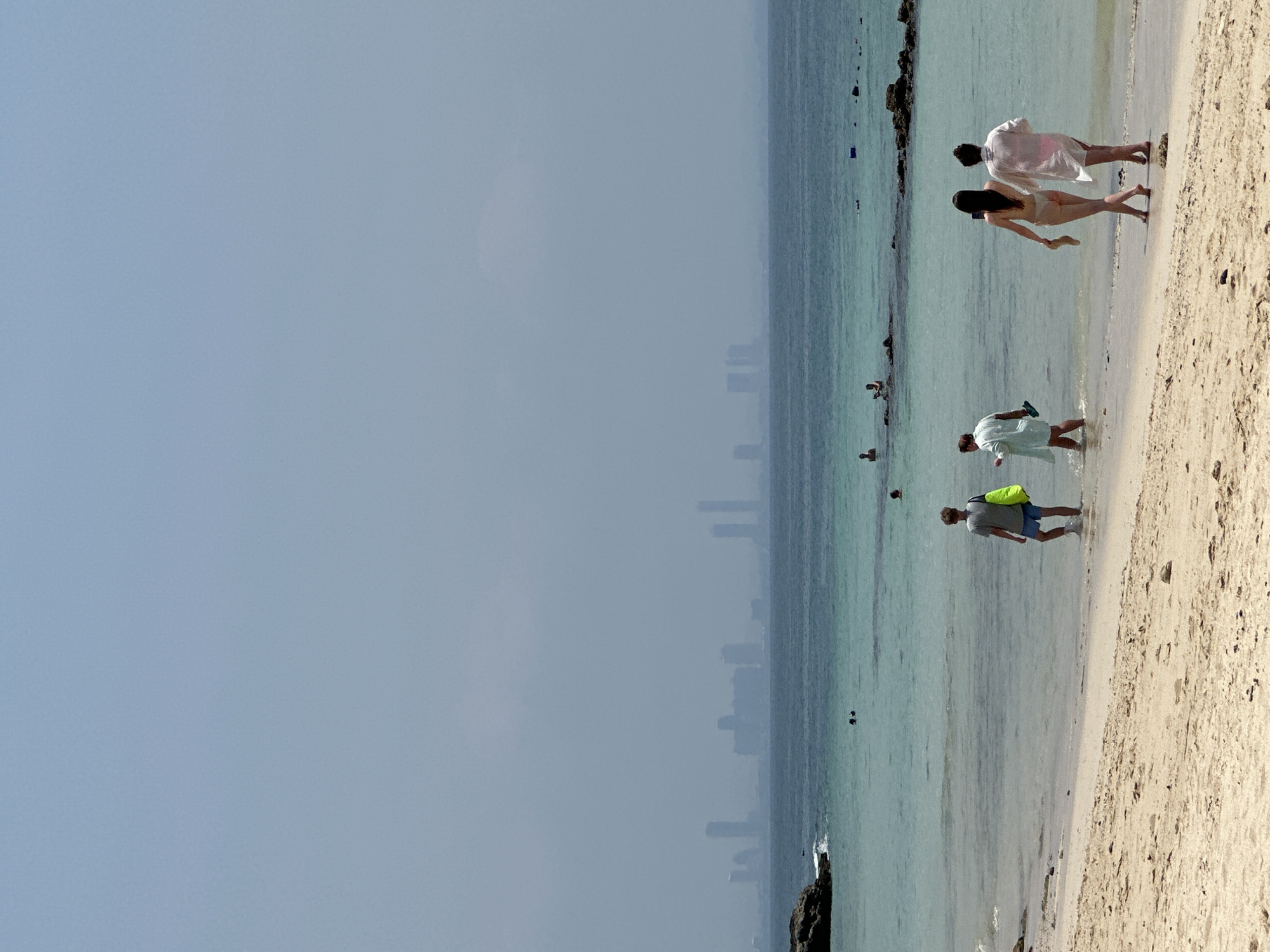 The image depicts a beach scene in Pattaya City, Thailand. The right side of the image features a sandy beach, with several people present. A couple is walking along the shoreline, while another person wades in the water. Another individual is seen walking along the beach further down. The water is a clear, turquoise color. In the distance, a faint skyline of buildings is visible across the water. The sky is overcast with a pale blue hue. Rocks are present along the water's edge.