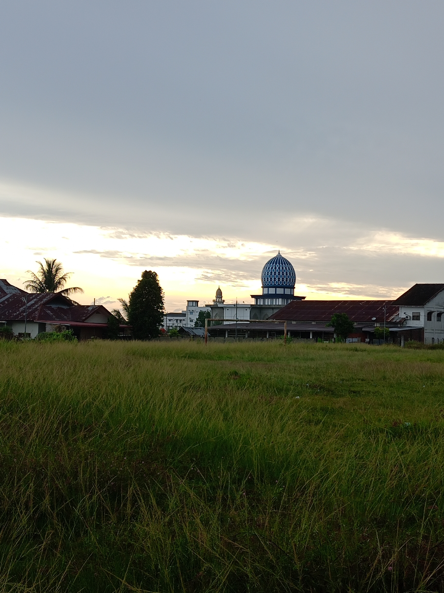 The photograph, taken in Geulanggang Baro, Indonesia, depicts a grassy field in the foreground with a cluster of buildings in the midground, and a partially cloudy sky in the background. The field is filled with tall green and brown grass. Behind the field, there are several buildings, including houses with reddish-brown roofs and a mosque with a blue and white striped dome and a golden spire. Other structures of note include an adjacent building with a long red roof, a two-story white building, and a single deciduous tree. The sky is filled with white and gray clouds. The sun is situated on the horizon, casting a bright glow on the scene.