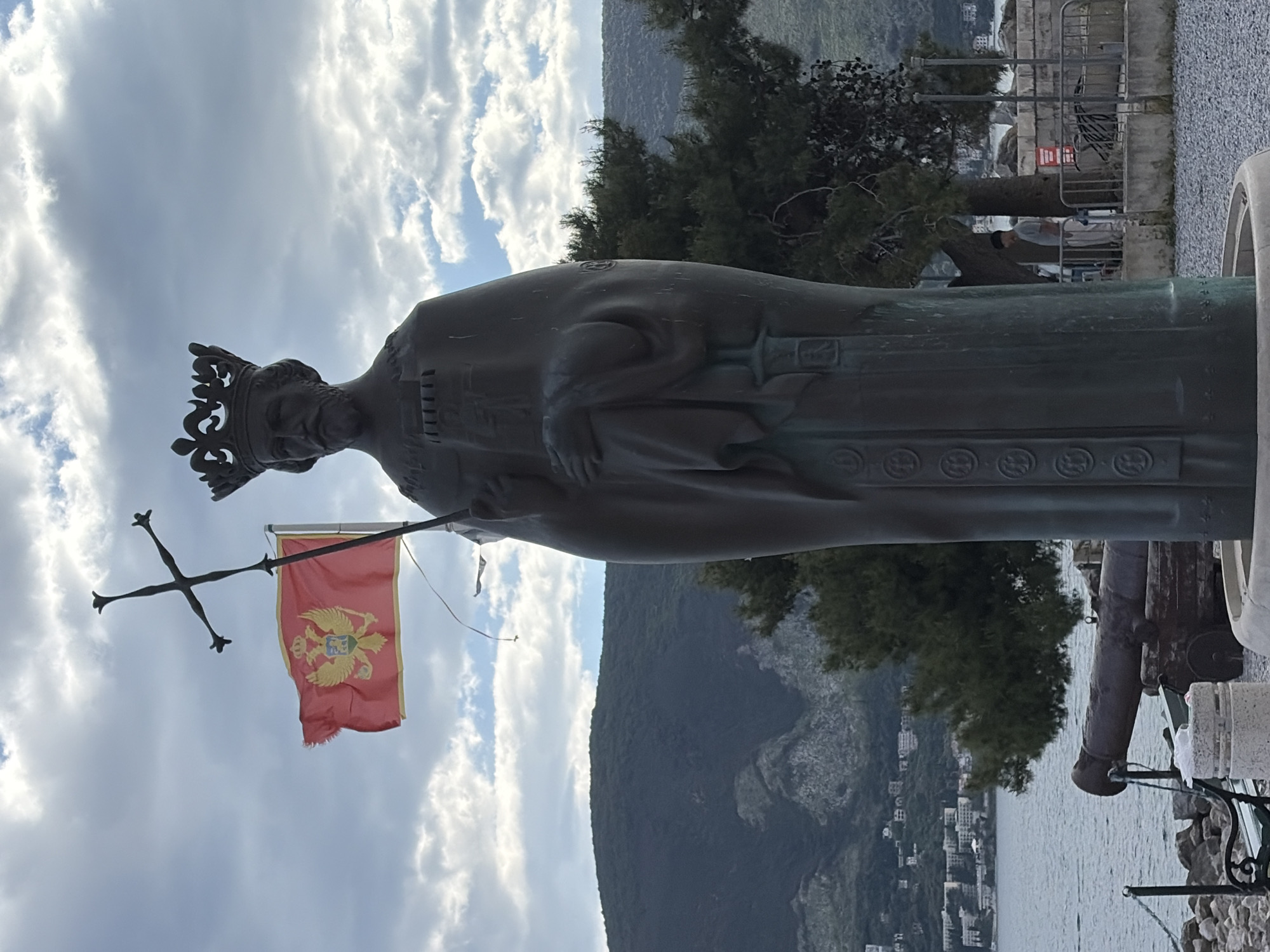 The image shows a bronze statue of a regal figure wearing a crown, holding a cross and a flag of Montenegro. The statue is located in Herceg Novi, Montenegro, with a view of the mountains, the bay, and buildings along the coast in the background. The statue is positioned on a stone base next to a cannon. Behind the statue is a green tree, and a metal fence. Above, the sky is filled with cloudy sky.