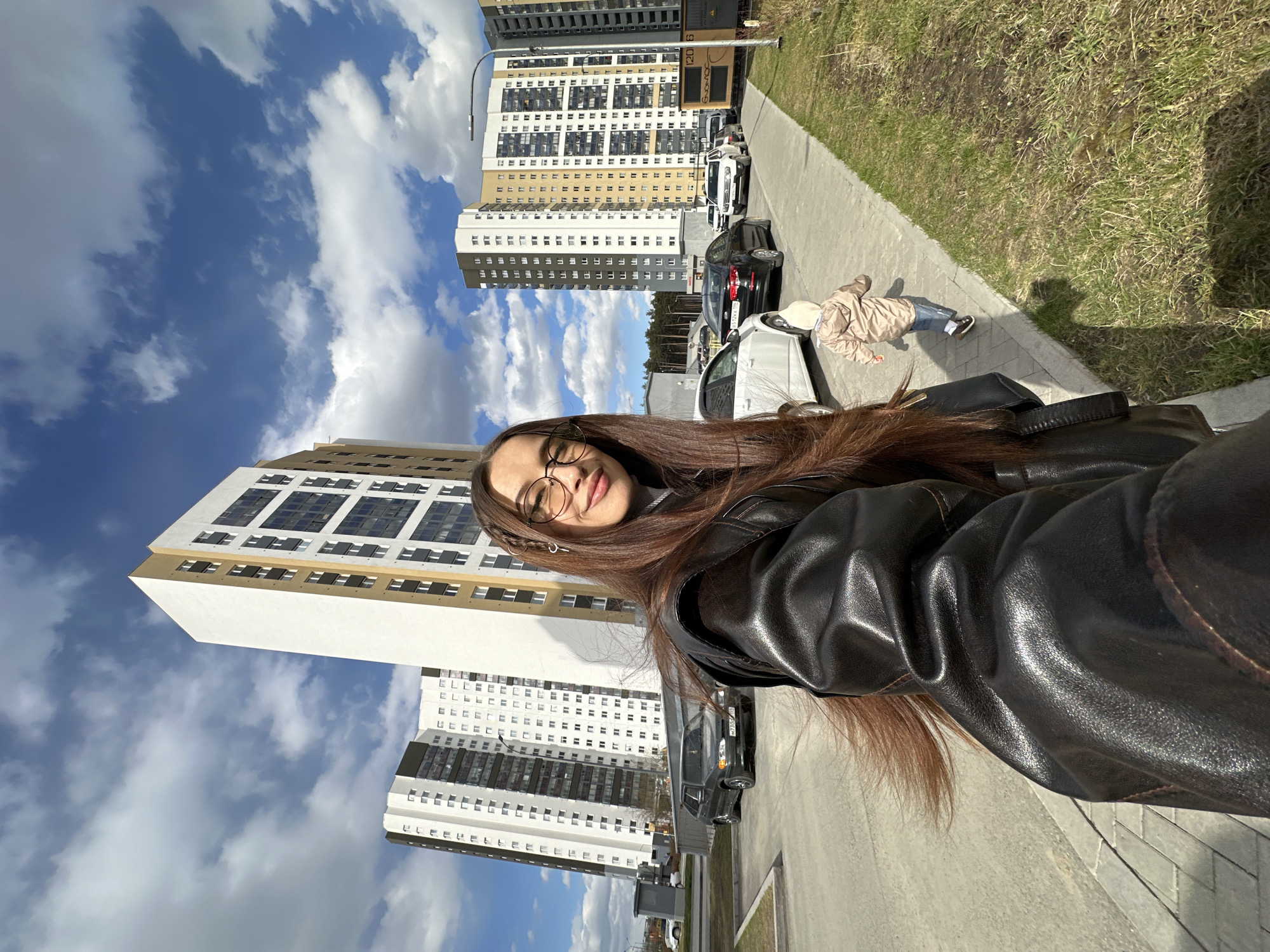 A selfie shows a young woman with long, straight dark brown hair, wearing round glasses and a dark (likely leather) jacket. She is smiling faintly at the camera. The image is rotated 90 degrees counter-clockwise, with the sky appearing on the left and the ground on the right of the current orientation.
In the background, several tall, modern apartment buildings dominate the urban landscape. These multi-story buildings feature a mix of white, grey, and light brown/beige facades with numerous windows. On one of the buildings, partial text "СОБКОМ" (SOBKOM) and numbers "120 6" are visible. Below and between the buildings, there are parked cars along a street. One car displays a partially visible Russian license plate, including "196 RUS," which corresponds to the Sverdlovsk Oblast, where Yekaterinburg is located.
To the right of the woman (as if the image were correctly oriented, i.e., in the mid-ground), a paved sidewalk runs alongside a strip of green grass. A small child, dressed in a light-colored jacket and jeans, is walking on the sidewalk with their back mostly to the camera. Strong sunlight suggests it's daytime, casting distinct shadows on the ground and from the child. The sky is bright blue with scattered white clouds, indicating clear weather. The overall scene depicts a sunny day in a modern residential area of Yekaterinburg, Russia.