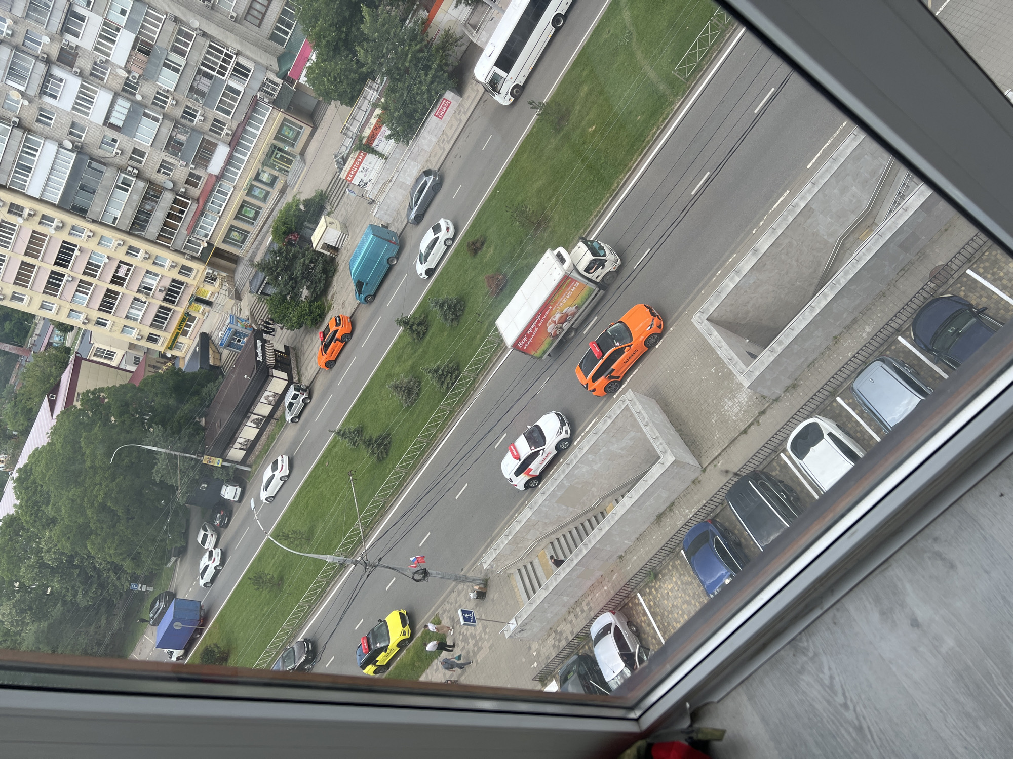 The image, taken from an elevated viewpoint, showcases a street scene in an urban environment. A multi-lane road runs horizontally across the frame, bordered by a grassy median strip. Several vehicles are in motion and parked. Among them are a white bus, a white delivery truck with advertising, an orange car, a yellow taxi, and several passenger cars of various colors including white, black, silver, and blue. On the sidewalk, three individuals are walking near a street sign and a blue recycling bin. Adjacent to the sidewalk is a concrete structure with stairs leading down, indicating an underpass or subway entrance. In the background, a large, multi-story apartment building with numerous windows dominates the skyline, alongside other smaller buildings and a dense area of trees. The lighting suggests it is daytime.