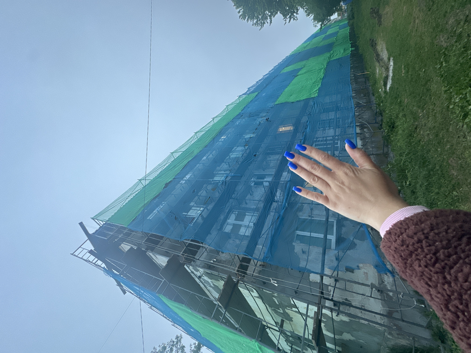 A low-angle, full shot shows a building covered in blue and green netting, indicating it is under renovation or repair. The building, which appears to be an apartment complex with multiple windows visible through the netting, is tall and angular, with scaffolding partially visible. In the foreground, a person's hand with bright blue painted fingernails is raised, obscuring part of the building. The hand is positioned in the lower right quadrant of the image. The environment around the building consists of a grassy area with some discolored patches, suggesting unkempt ground. The sky is overcast and a light color, contributing to a muted atmosphere. The scene is set in the city of Vilyuchinsk, Russia, a location known for its naval bases and proximity to volcanoes, though no specific landmarks in the image identify it as such beyond the provided context. The overall impression is one of ongoing construction work in a residential urban setting.