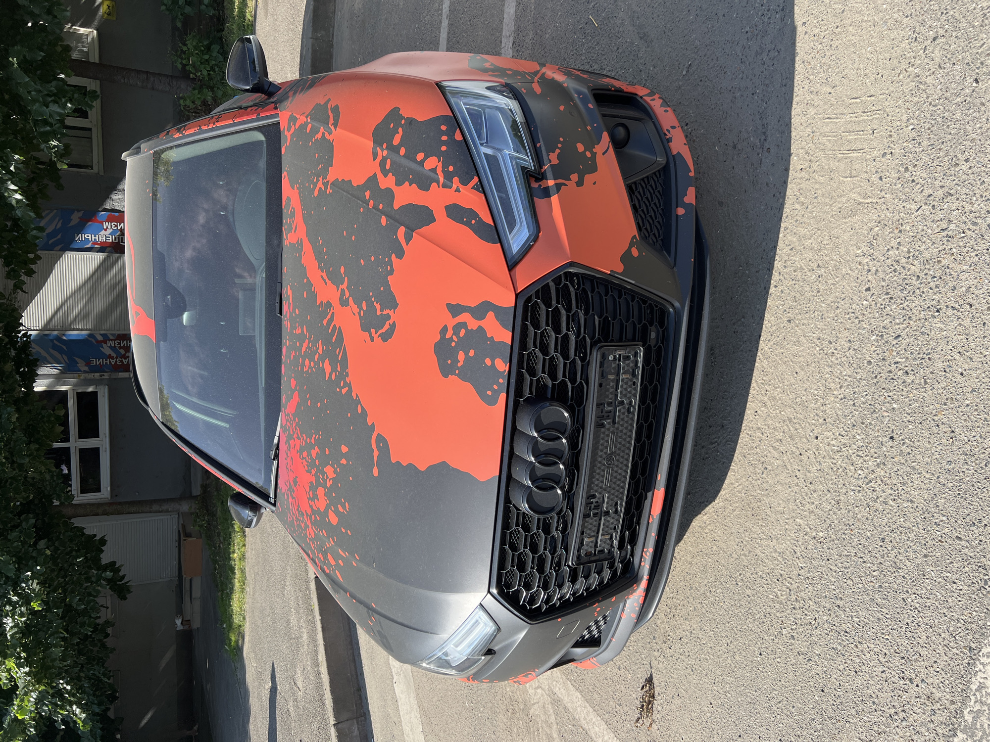 An Audi A4 Avant wagon with a matte grey and bright orange splatter-paint wrap is parked on asphalt in front of a building with trees and signage. The car is facing forward, with its headlights and grille visible. The building appears to be a commercial establishment with windows and a shuttered entrance. The environment suggests a sunny day in Krasnodar, Russia.