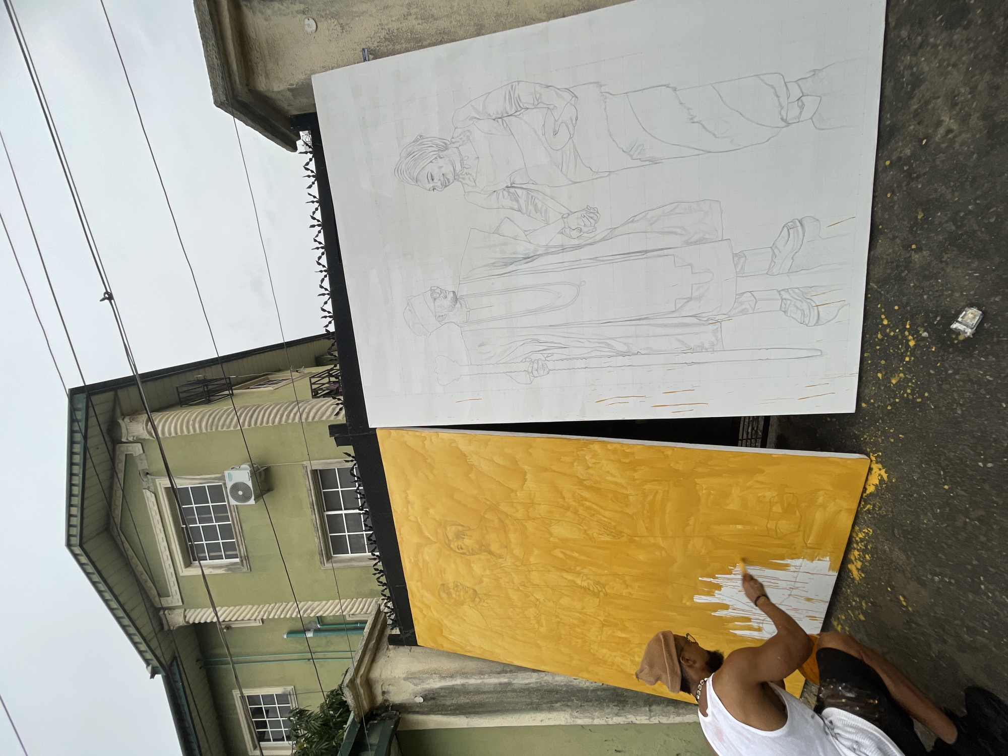 A man is painting outdoors in Ado, Nigeria. He is sitting on a paved surface beside a green building with windows and an air conditioner unit. The man is wearing a white tank top, shorts, a hat, and a beaded necklace. He is applying yellow paint to a large canvas with a sketched outline of two figures. Above the yellow canvas is another canvas with a similar sketch, but it is unpainted and white. Splatters of yellow paint are visible on the ground near the canvases.