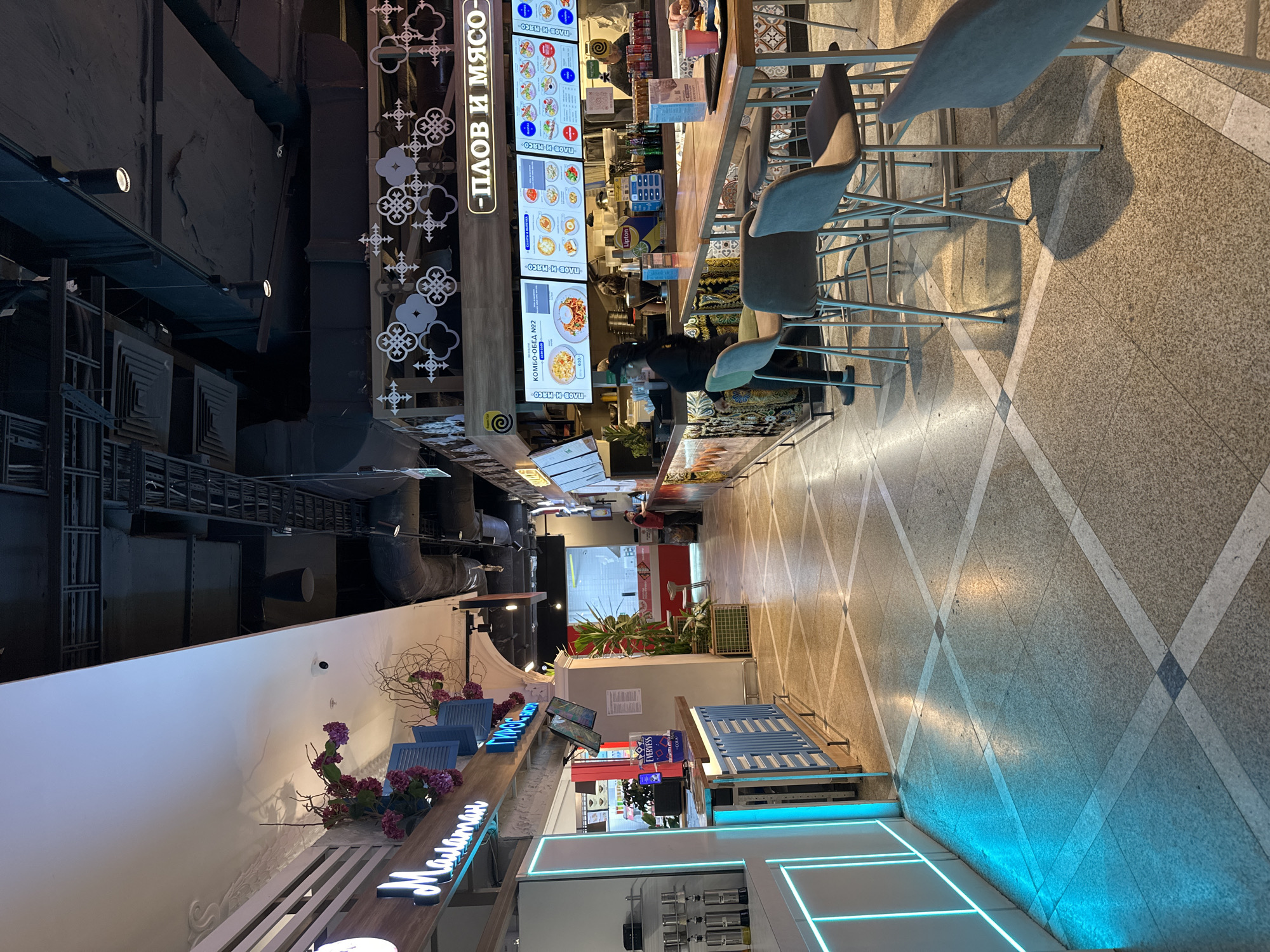 This image depicts an indoor food court or shopping mall area in Yekaterinburg, Russia, featuring multiple food establishments and a contemporary design. The central focus is a food stall named "ПЛОВ И МЯСО" (Plov i Myaso), which translates to "Pilaf and Meat," easily identifiable by its prominent vertical sign adorned with decorative patterns.
At the "ПЛОВ И МЯСО" stall, several digital menu boards display food items, with visible text including "КОМБО-ОБЕД №2" (Combo Meal №2) and "САЛАТЫ И ВЫПЕЧКА" (Salads and Baked Goods). A person in a black cap is seen behind the counter, attending to the stall, while another individual is partially visible further along the counter, possibly a patron. A row of grey, modern bar stools with light metal frames lines the counter, providing seating for diners. Brands like Lipton and Evervess are discernible on products at the counter.
The floor throughout the area is a light-colored speckled tile with a distinctive pattern of diagonal white lines forming a grid. Further down the concourse, another shop's signage, featuring blue neon letters that partially read "Мариман" (Mariman) and is decorated with purple floral arrangements and wooden elements, adds to the aesthetic. Blue slatted structures and benches are also visible in this area, complementing the glowing blue neon strips integrated into the floor design. Overhead, the ceiling reveals an industrial aesthetic with exposed dark pipes, ducts, and structural elements. Another shop sign, "Галатан" (Galatan), is partially visible in the wider shot. The overall scene suggests a vibrant, modern commercial environment.
