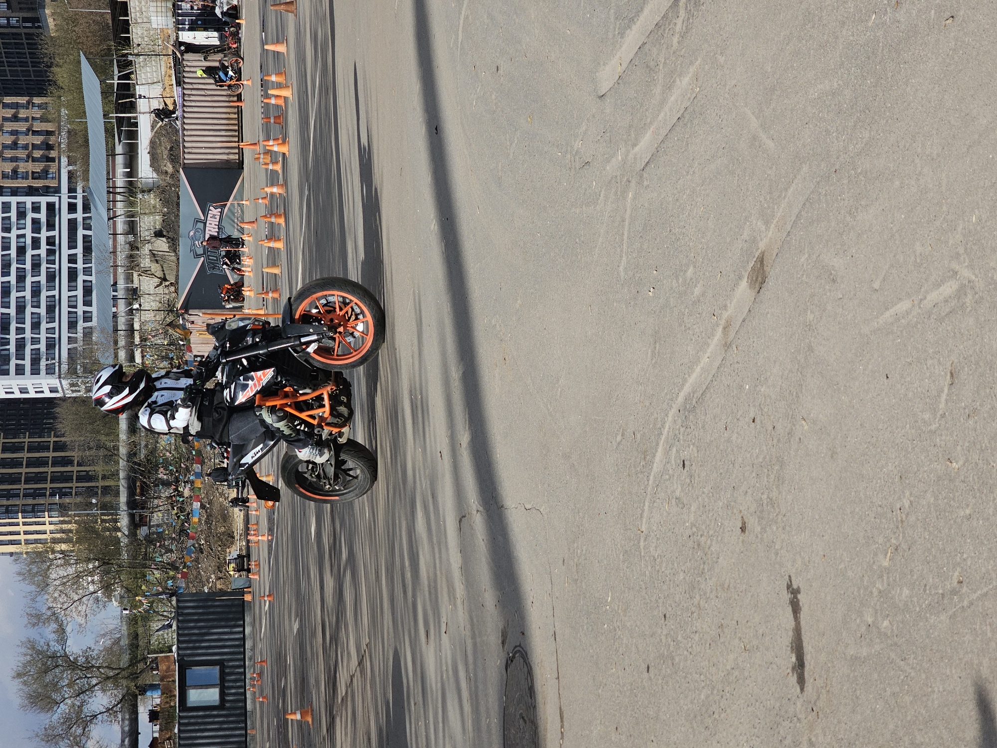An outdoor, wide-angle, medium shot captures a motorcycle rider performing a maneuver on a paved surface, set against an urban backdrop.
In the foreground and midground, a person wearing a white and black helmet, a white and black protective jacket, gloves, and dark pants is seated on a black and orange KTM sports motorcycle. The motorcycle's front wheel is slightly elevated from the ground, indicating a possible wheelie or balancing exercise. The rider and motorcycle are positioned on an asphalt surface, which is marked by numerous orange traffic cones arranged in rows, defining a training course or event area.
Long, distinct shadows are cast across the pavement from the left side of the frame, suggesting a sunny day. A manhole cover is visible on the asphalt to the right. In the background, a large black banner displays the white text "Y HACK TRACK". Behind the banner, several corrugated metal structures, resembling shipping containers, are visible. Further in the distance, a mix of trees, some with bare branches and others with green foliage, stands before a cluster of multi-story buildings. These include a prominent white building with many windows on the far left and other brown and grey structures.
The scene depicts a dynamic moment of motorcycle training or a performance event during daylight hours under clear skies, in what appears to be a dedicated urban or semi-industrial practice area.
