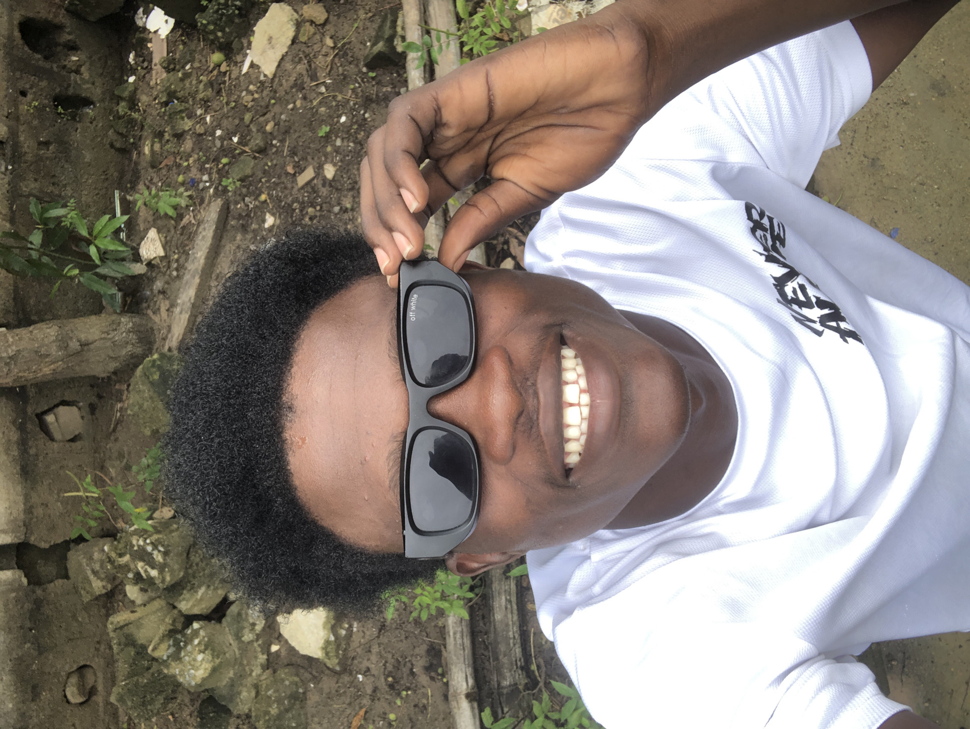 A young Black male is pictured from the chest up, outdoors, with a slight upward angle. He is wearing a white t-shirt with black lettering on the left side of the chest, which appears to read "NEVER ALL". He has short, dark, curly hair and is wearing dark sunglasses with "off white" printed on the left lens. His right hand is raised, and he is holding the sunglasses on the right temple with his index finger and thumb. He is smiling broadly, revealing his teeth. The background consists of a grey, weathered concrete wall, some broken bricks, dirt, and green foliage. The scene is consistent with an outdoor location in Ado, Nigeria, possibly a residential area or an alleyway, given the worn appearance of the surroundings.