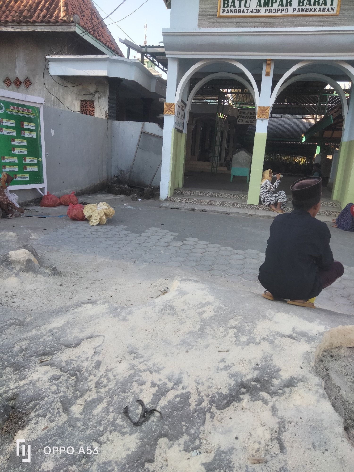 The image depicts an outdoor scene near what appears to be a religious or communal building, possibly in Badung, Indonesia. The architecture suggests a traditional Indonesian style with a red-tiled roof visible on the left and arched structures with white columns and gold accents on the right, forming an entrance. Above the arches, a sign reads "BATU AMPAR BARAT PANGSBATHOK PROHPO PAMEKASAN," indicating the location or name of the place.
In the foreground and midground, the ground is paved with hexagonal concrete tiles and has areas of loose sand and dirt. Several people are present:
- A person in the foreground, seen from behind, is crouched on the ground, wearing a dark shirt, dark pants, and a dark cap. They have yellow sandals on.
- To the left, a woman in a light-colored headscarf and a patterned dress is seated on the steps of the building. She is holding a bottle and appears to be drinking.
- Further to the left, a person is seated on the ground, partially obscured by a green sign board.
- Another person in the far right background is also seated, wearing a patterned garment and a headscarf.
Objects in the scene include:
- A large green sign board on the left with text and images, likely displaying organizational information or a directory.
- Several red plastic bags and a large, light-colored, possibly fabric, bundle are placed on the ground near the sign board.
- Debris, including small rocks and what looks like dried vegetation, is scattered on the ground.
- A dark, coiled object, possibly a snake skin or a dead snake, is visible on the sandy ground in the lower center of the image.
The lighting suggests it is daytime, with sunlight casting some shadows. The overall environment appears to be an open area adjacent to a building, possibly a mosque or a public gathering space. The presence of sand, debris, and the people's attire suggest a warm climate. The watermark "OPPO A53" is present in the bottom left corner.