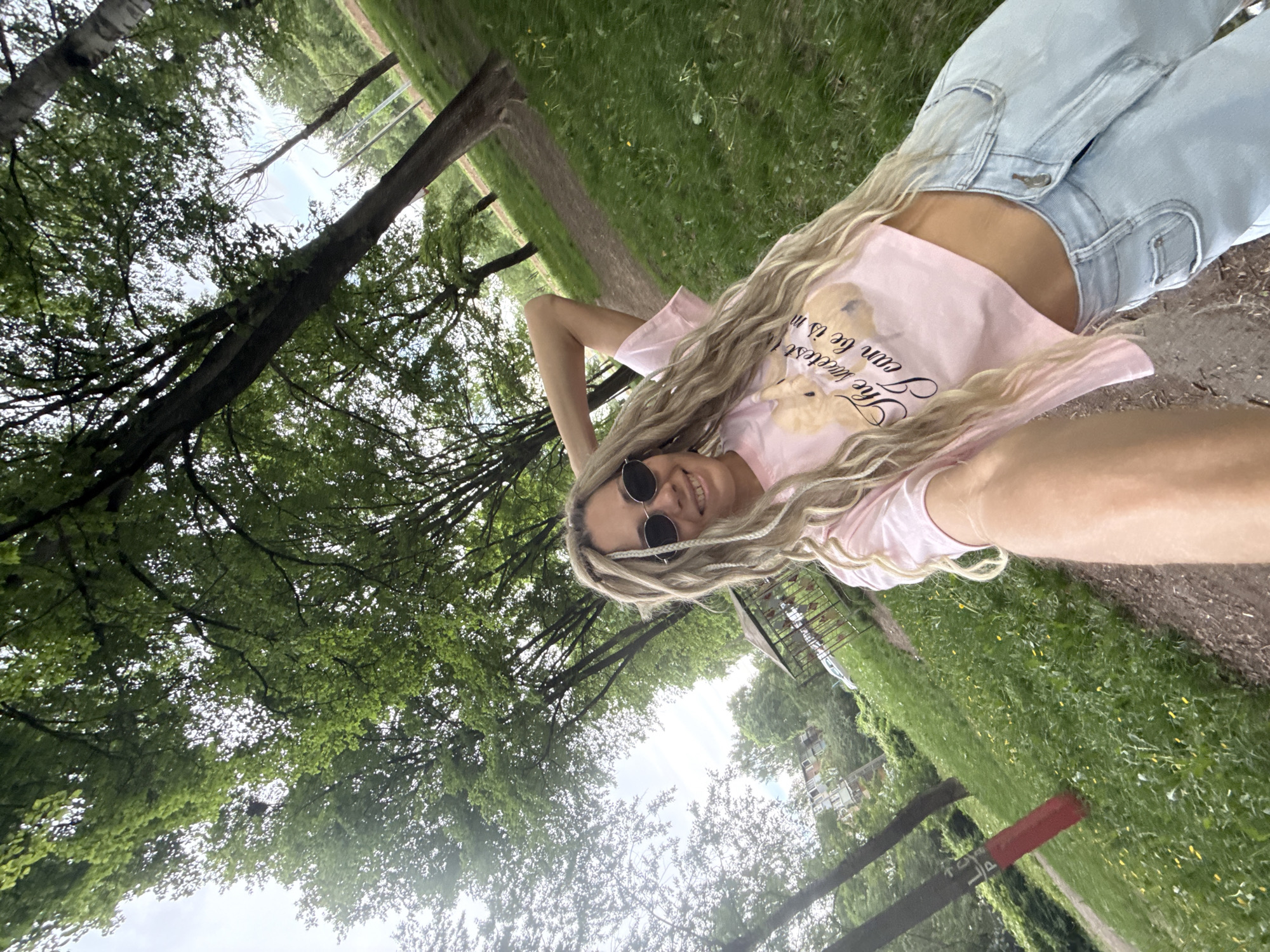 A young woman is lying on a concrete surface in a park-like setting in Rusanovo, Russia. She is wearing light blue jeans, a pink t-shirt with a teddy bear graphic, and round sunglasses. Her long blonde hair is styled in braids or dreadlocks. The ground around her is covered in grass and trees. A fence or barrier is visible in the background. The camera angle is low, looking up at the woman and the trees.