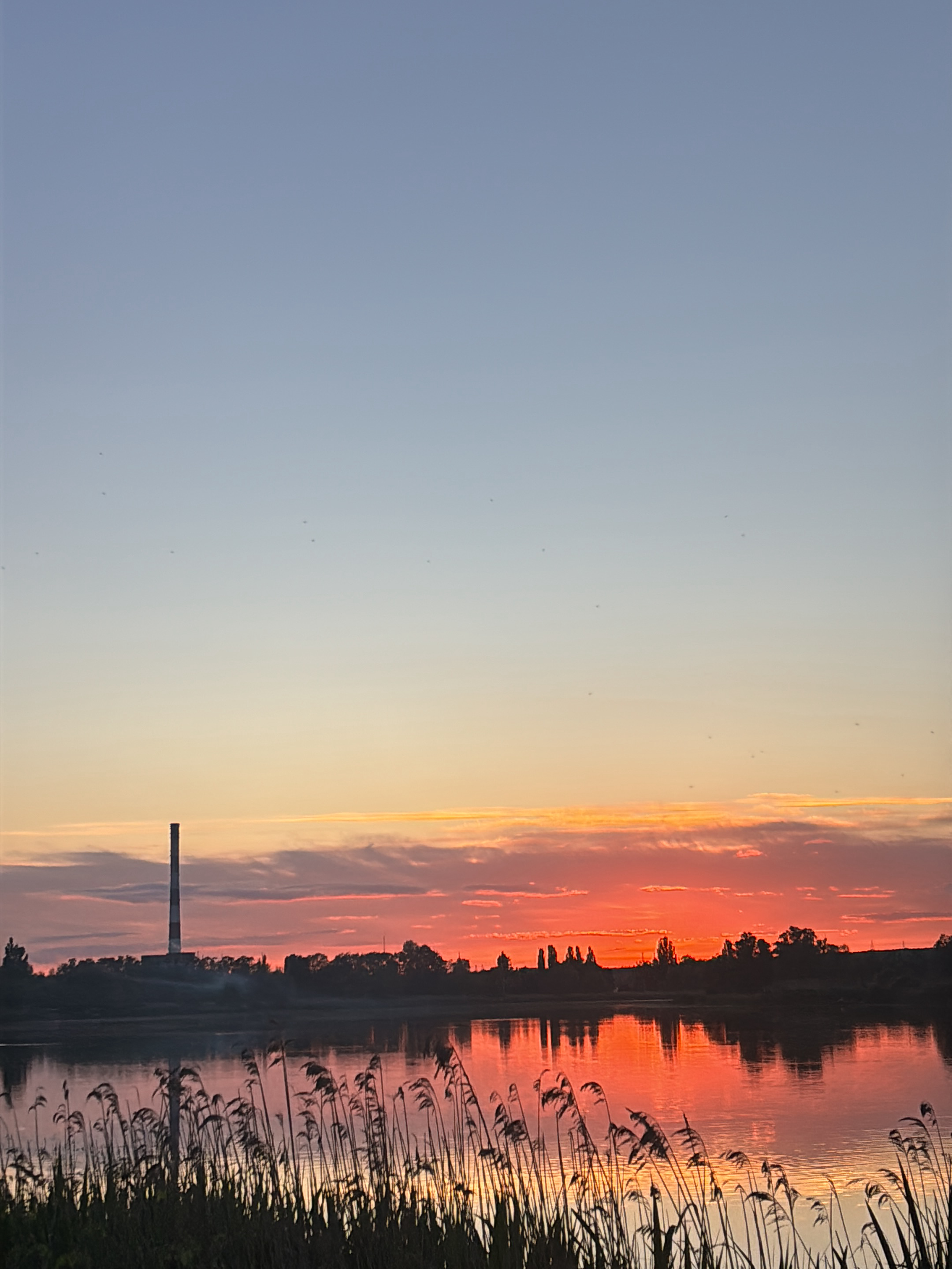 The image depicts a sunset over a body of water, with reeds in the foreground. A tall, industrial chimney stands on the left side of the horizon. The sky transitions from a pale blue at the top to shades of orange and red near the horizon. The water reflects the colors of the sunset. The location is identified as Pisochyn, Ukraine. The scene contains no visible people.