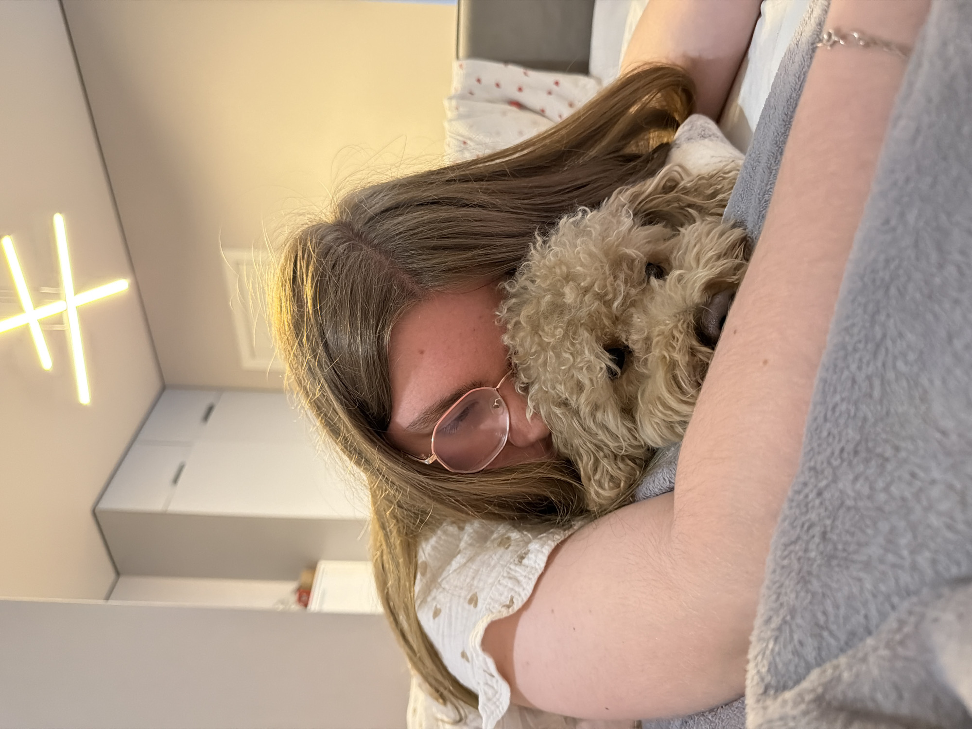 An indoor scene depicts a person lying prone, embracing a small, light brown, curly-haired dog, located in Perm, Russia. The individual, possessing long, light brown hair, is wearing a light-colored top adorned with small heart patterns and distinctive rose-tinted, possibly octagonal, eyeglasses. A silver-colored link bracelet is present on the person's right wrist. The dog, a poodle-type breed, is nestled closely, its head resting on the person's arm, with its face directed towards the viewer. A grey, textured blanket occupies the immediate foreground. The background reveals a modern room with light-colored walls, a partially visible white bed frame, and white bedding, some of which has faint red patterns. Overhead, a contemporary ceiling light fixture consisting of four linear light bars arranged in a cross shape illuminates the space. A tall, white cabinet with dark horizontal handles is visible on the left side of the room.