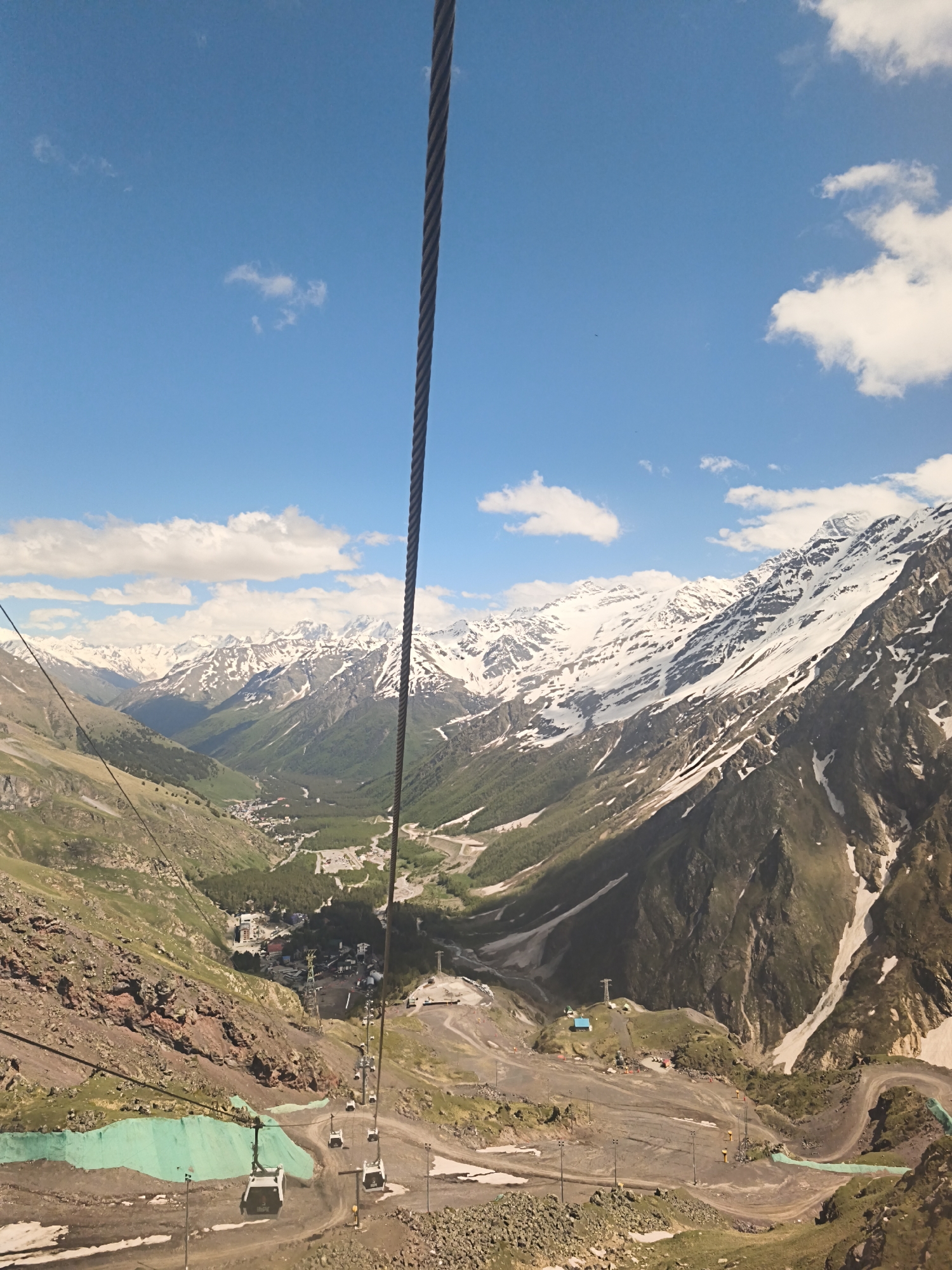 The image is a high-angle view of a mountainous landscape near Terskol, Russia, likely taken from a cable car. A thick, dark cable runs vertically down the center-right of the frame. Below, a valley is visible, containing a small town with buildings and roads. Steep, rocky slopes with patches of green vegetation flank the valley. Further in the distance, snow-capped mountains form a dramatic backdrop under a bright blue sky with scattered white clouds. Several cable car cabins are visible descending the mountainside. The terrain in the foreground is rocky and uneven, with some areas covered by green geotextile fabric and patches of lingering snow. The overall scene depicts a rugged, high-altitude environment with infrastructure for tourism or transport.
