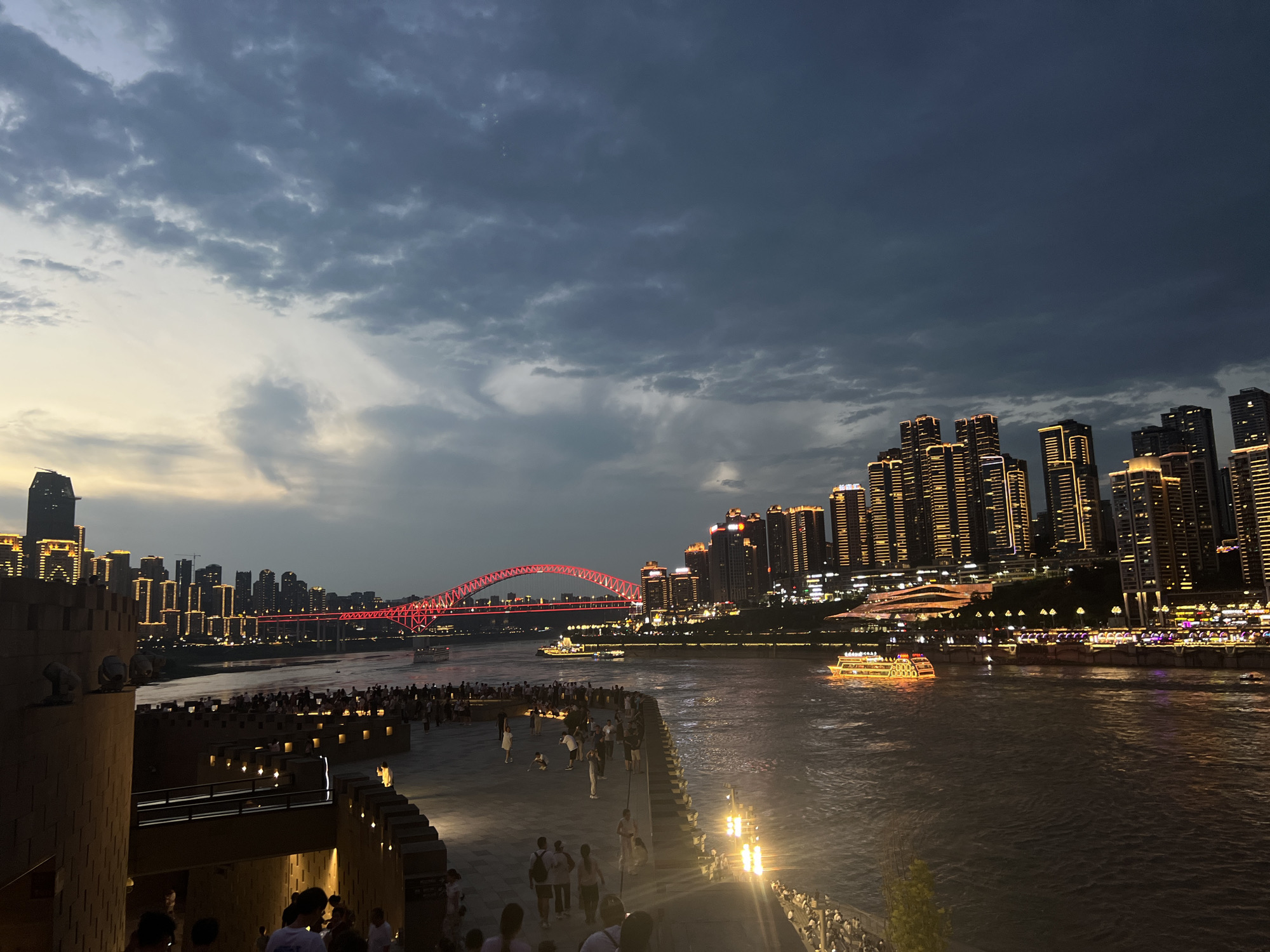 The image depicts a twilight scene in the Yuzhong District of China, featuring a riverfront cityscape with a bridge and numerous buildings illuminated by artificial lights. In the foreground, a stone structure resembling a fortress with spotlights is visible, and a paved walkway is populated by many people. Several individuals are walking and standing on the walkway. The river, which spans the middle ground, has a notable presence of illuminated boats, including a large yellow vessel with glowing windows and a white boat. Spanning the river is a red-arched bridge, also brightly lit. Across the river, a dense collection of modern high-rise buildings forms the city skyline, their windows emitting warm yellow light. The sky above is filled with dramatic, dark clouds.