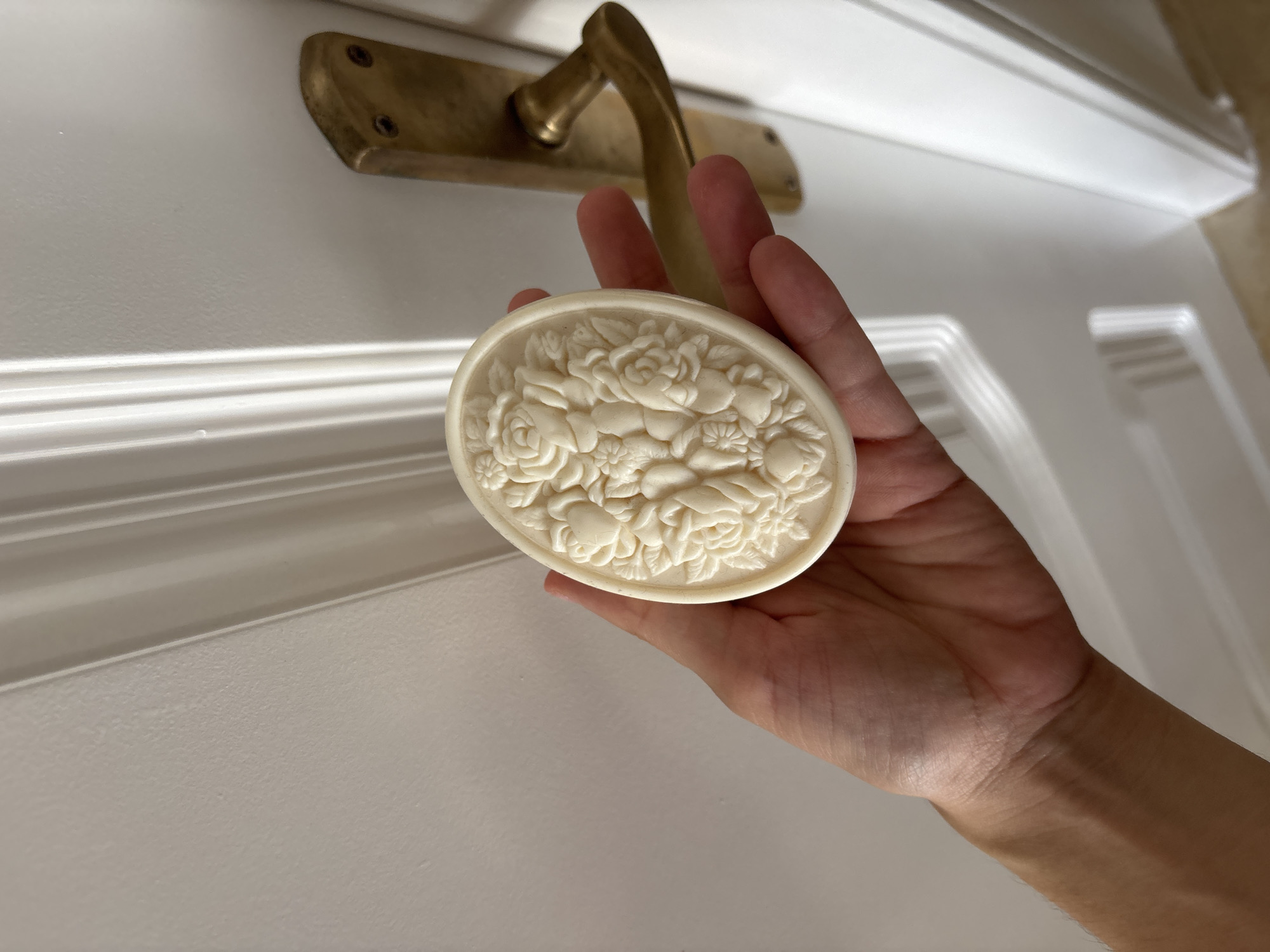 A hand holds an oval-shaped, intricately carved soap with a floral design of roses and other flowers. The soap is a light cream color and has a matte finish. The hand is lightly tanned, with visible veins. Behind the hand and soap is a white door with horizontal molding. Above the door, a brass door handle is visible, showing signs of age and slight tarnish. The background is softly lit, suggesting an indoor setting. The overall impression is one of delicate artistry and domesticity. The image does not contain any visible text. The location context is Orihuela, Spain.