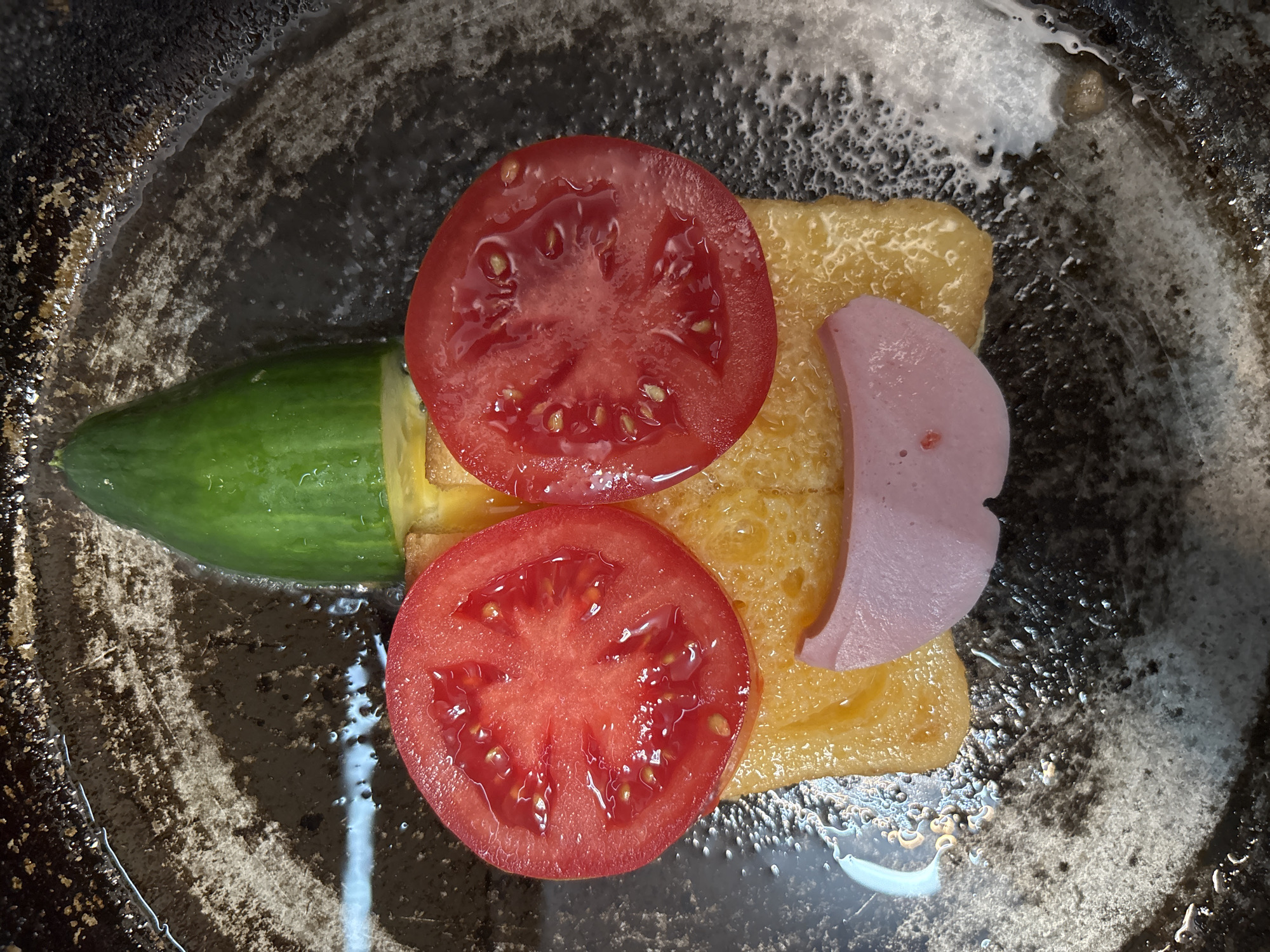The image shows a plate containing a cucumber, two slices of tomato, a slice of tamagoyaki (Japanese rolled omelet), and a slice of ham. The plate has a dark, mottled appearance. The food items are arranged on the plate in a somewhat abstract manner. There is no discernible connection to Izhevsk, Russia, or any identifiable people, actions, or interactions.