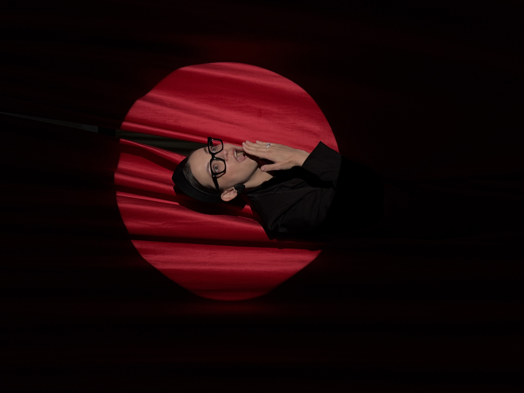A person with short dark hair, wearing glasses, a black shirt, and a head covering, appears within a circular spotlight on a red, draped surface. They are looking up with their mouth open and their right hand raised to their mouth. The background is dark. The photograph does not provide enough information to determine if the location is in the city of Chaikovsky, Russia.