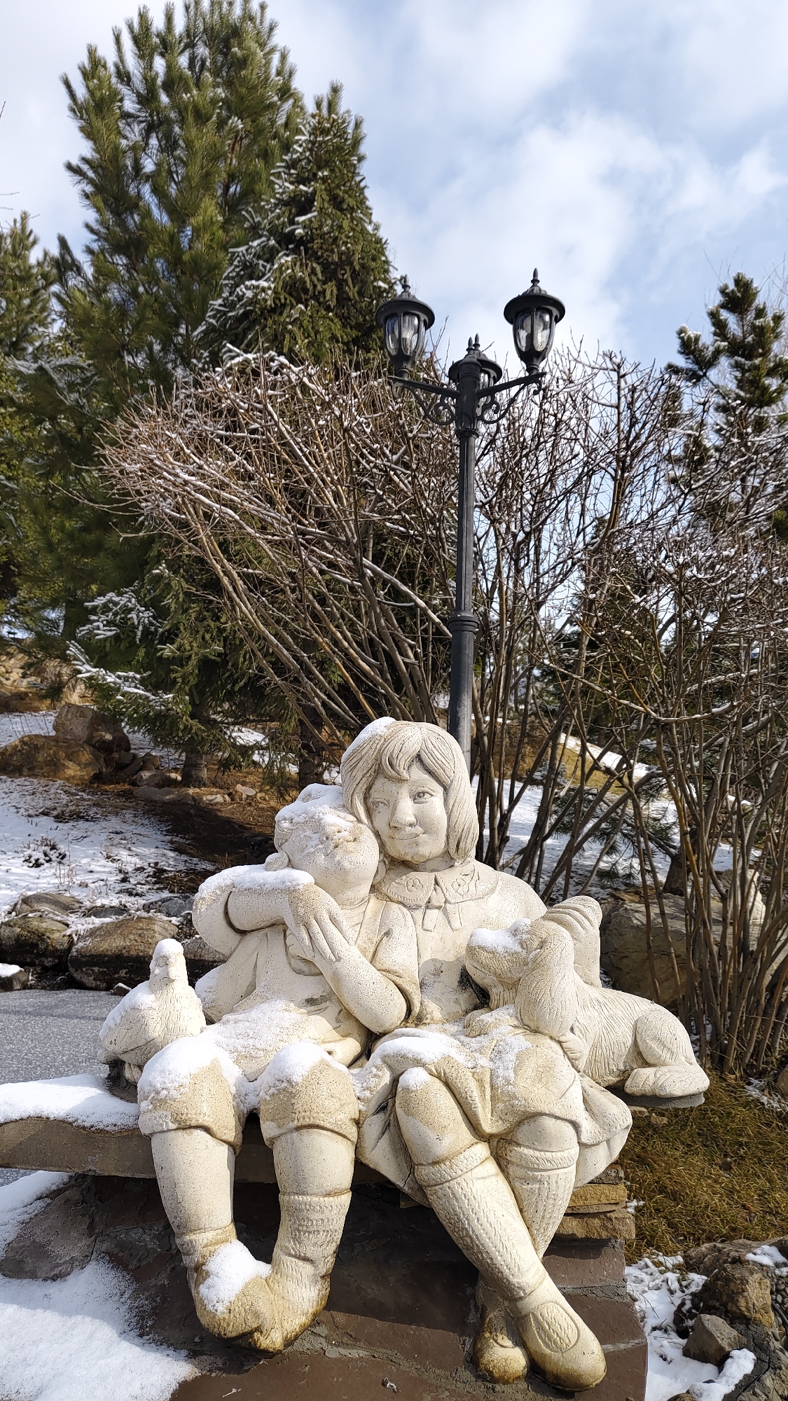 The image shows a stone sculpture of a young girl with a child and a dog, all covered with snow. The sculpture is seated on a stone bench. Behind the sculpture, there is a black lamppost with three lamps, bare bushes and evergreen trees. The ground is partially covered with snow, with visible rocks and patches of grass. The sky is visible in the background. The location is in Krasnoyarsk, Russia.