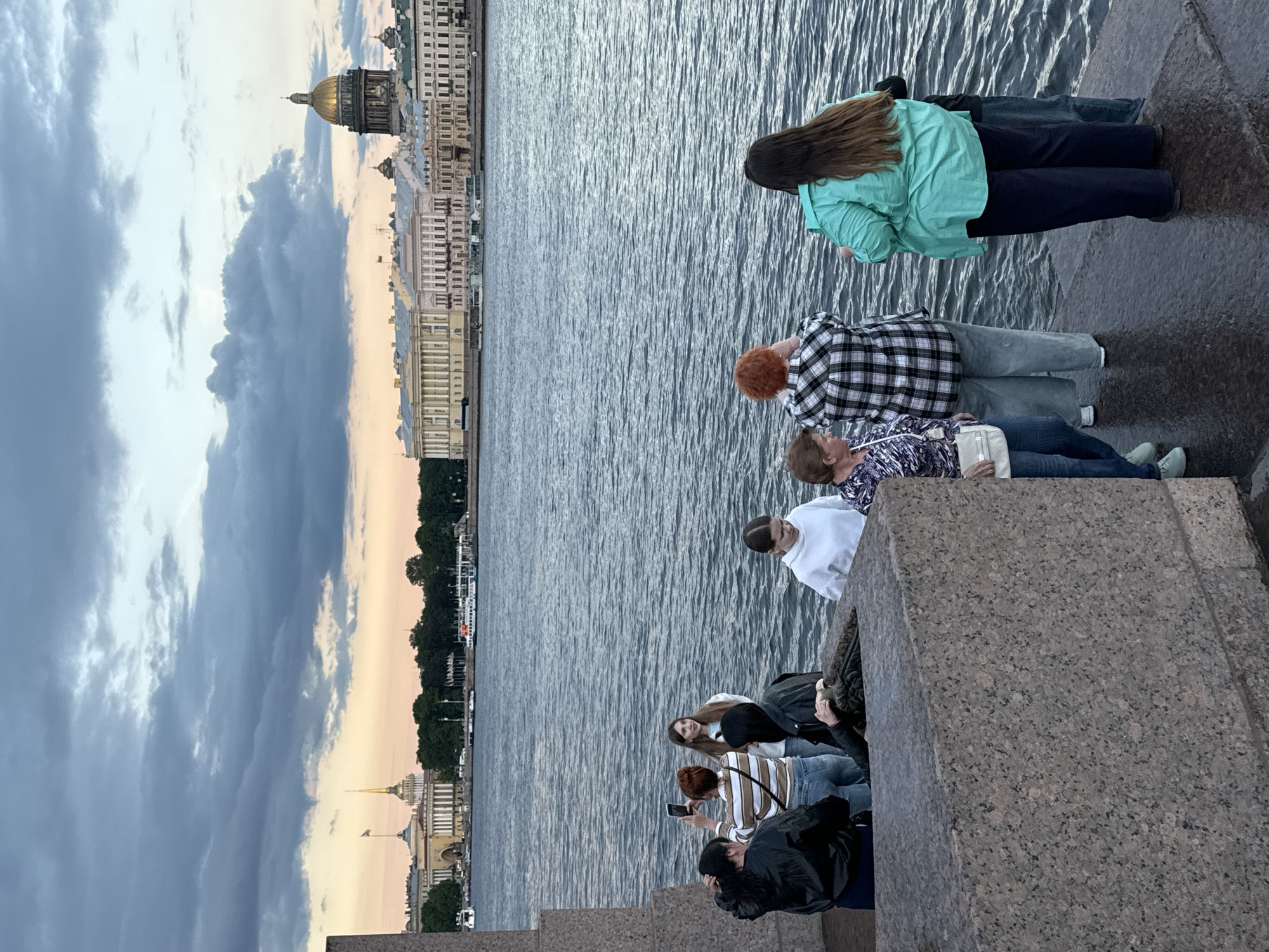 A group of six people are gathered by the water's edge in Saint Petersburg, Russia. The sky is overcast with dark, dramatic clouds, and the water is calm. In the background, across the Neva River, a row of grand, classical buildings lines the embankment. Notable structures include a building with a spire and flag, and further to the right, a large building topped with a golden dome, likely St. Isaac's Cathedral. Several boats are visible on the water.
In the foreground, a large, rough-hewn granite structure forms a barrier. Five individuals are on the paved embankment behind this barrier, facing the water.
On the left, two women are standing. One, wearing a black jacket, is holding a phone and appears to be taking a photo or video of the person in front of her. This second woman, with red hair, is wearing a striped top and jeans and is looking towards the water. To her right, a young woman with long brown hair is standing and looking towards the right.
Further to the right, another young woman in a white hoodie and jeans is looking down. Next to her, a woman with red hair is wearing a black and white plaid shirt and grey pants, facing away from the camera, looking out at the water. To her right, a woman with long brown hair is wearing a bright teal shirt and dark pants, also looking out at the water. A sixth person, wearing a dark jacket and blue jeans, is partially visible behind her, also looking towards the water.
The overall scene depicts a group of tourists or locals enjoying the waterfront views of Saint Petersburg.