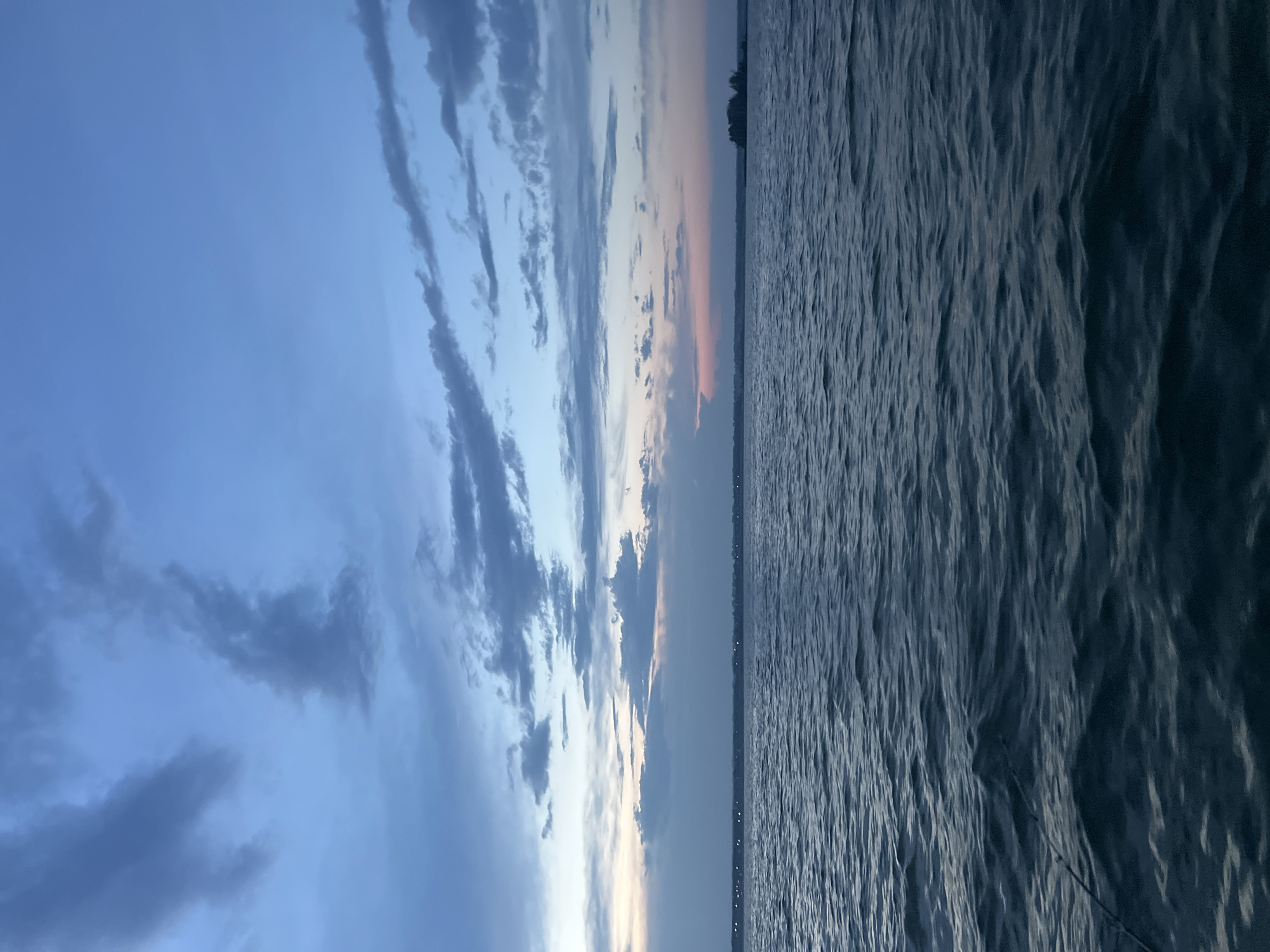 The image depicts a serene body of water, likely a lake or a wide river, under a twilight sky. The water is dark blue with gentle ripples, suggesting a calm atmosphere. The sky is a gradient of blues, transitioning from a deep indigo at the top to lighter hues near the horizon, where faint traces of pink and orange are visible, indicating the setting sun. Wispy clouds are scattered across the sky, some appearing dark and textured, while others are softer and more diffuse.
In the distance, a faint line of lights suggests a shoreline with some form of settlement or buildings. To the far right, a small, dark island or peninsula with several trees is discernible. In the foreground, a fishing rod is partially visible, extending from the lower left corner of the frame and resting on the water's surface, implying an individual is fishing.
The overall scene evokes a sense of tranquility and quietude. Given the context of Novopichugovo, Russia, this could be a view of a popular fishing spot or a scenic area along the Yenisei River, which flows near the region. The time of day is likely dusk or early evening.