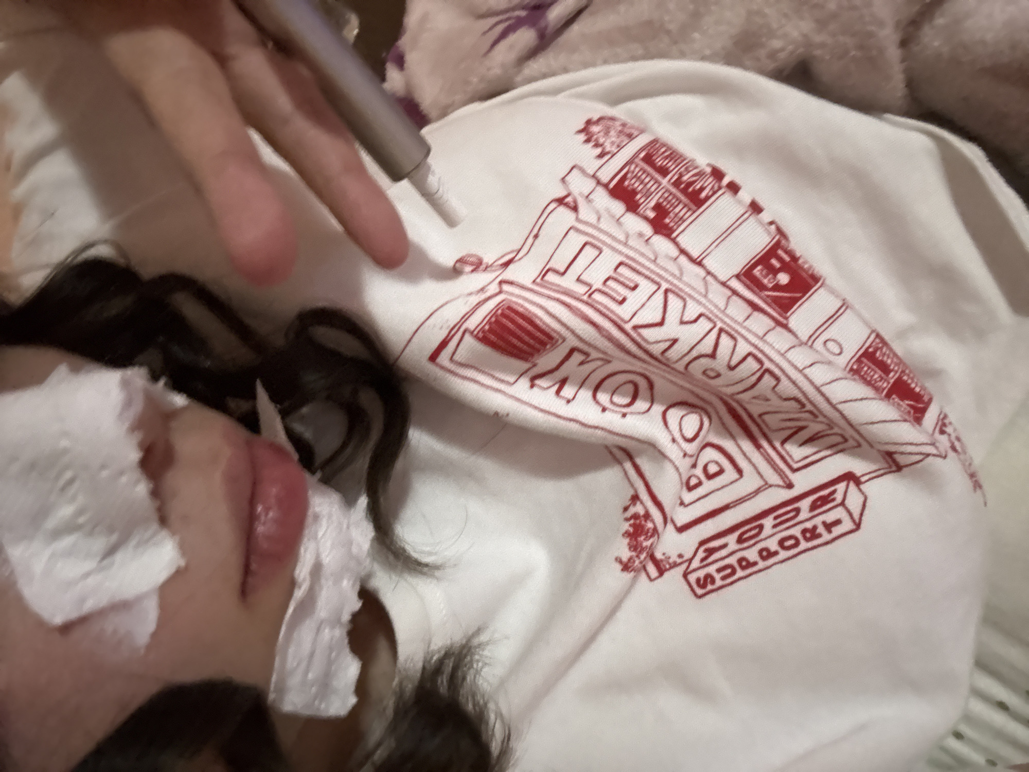 A person with dark hair and facial coverings is lying down, wearing a white t-shirt with a red graphic that includes the words "Book Market" and "Your Support." A hand holding a silver, pen-like object is positioned above the t-shirt. The location in Garmen, Bulgaria, is not discernible from the image.