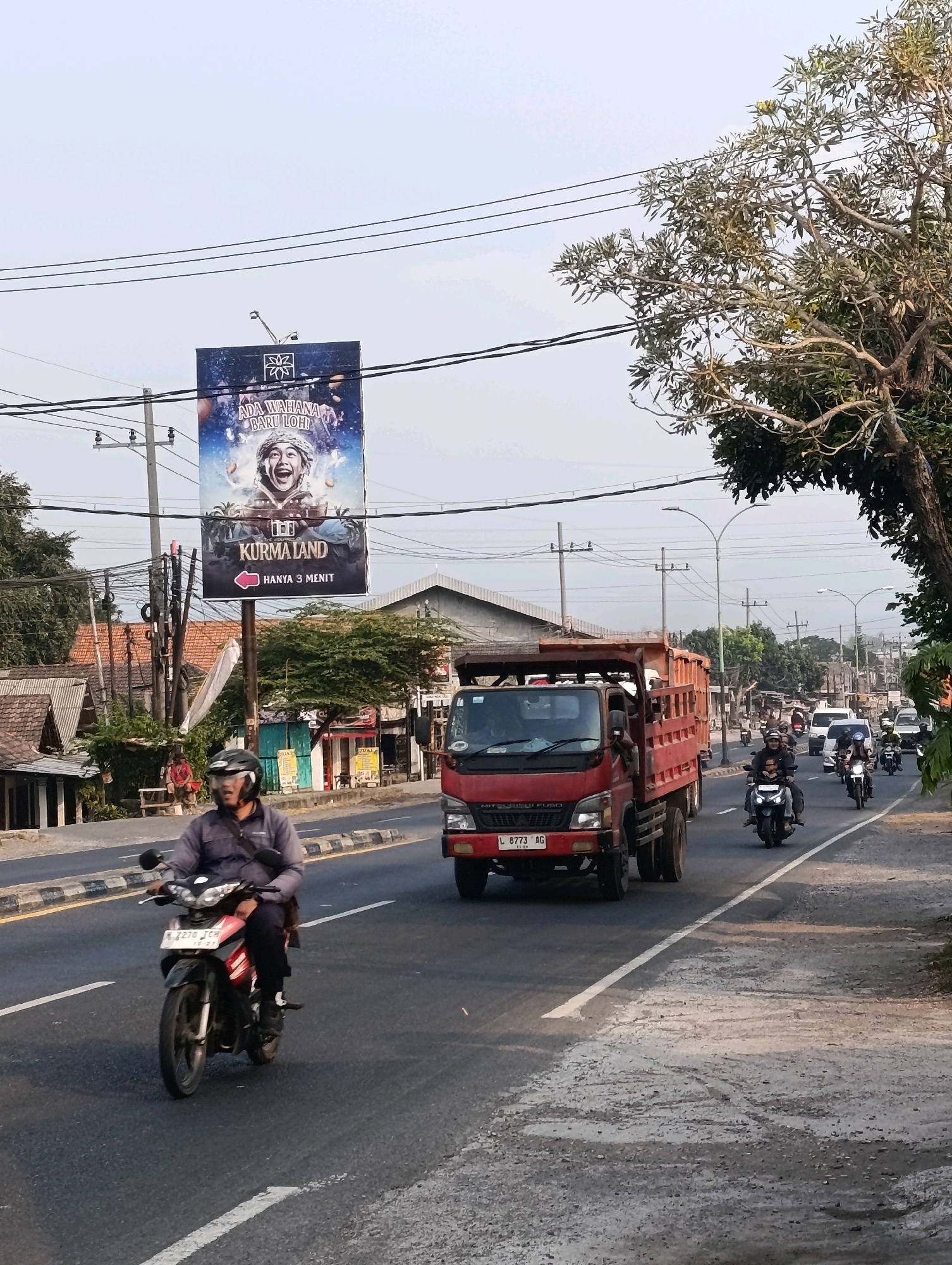 The image depicts a street scene in Karangsono, Indonesia, under a clear sky. A large billboard advertising "Kurma Land" with the tagline "Ada Wahana Baru Loh! Hanya 3 Menit" is prominently displayed above the road. In the foreground, a person is riding a motorcycle, wearing a dark jacket and a helmet. Behind them, a red truck is driving on the same road, followed by several other motorcycles and a white car. The road is paved and marked with white lines. On the left side of the road, there are several buildings with traditional Indonesian architecture, including houses with tiled roofs. Trees line the right side of the road, some with sparse foliage. The overall impression is a typical urban or suburban street with ongoing traffic and commercial advertising.