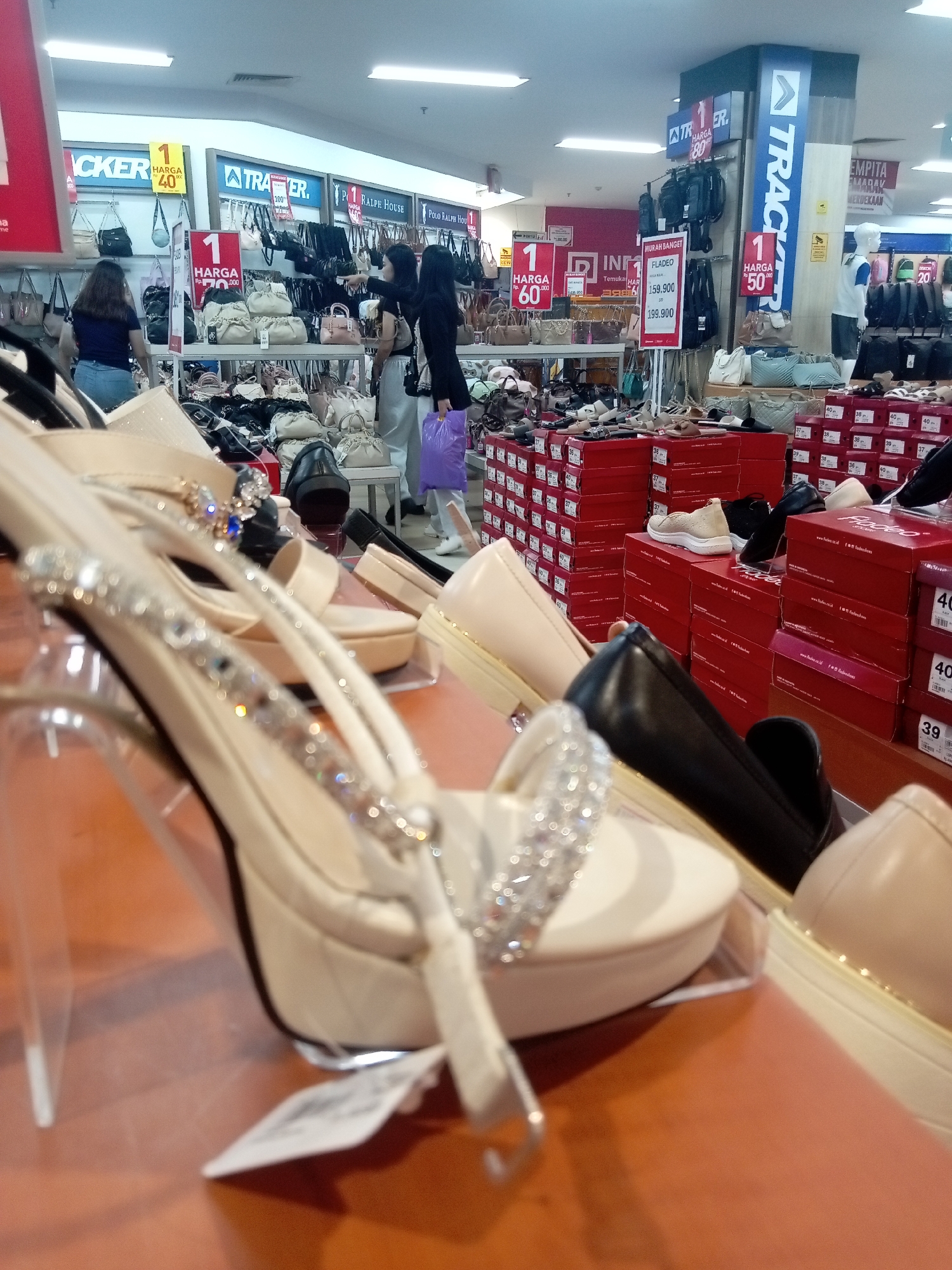 The image depicts the interior of a department store, likely in Batam, Indonesia, judging by the signage and language used. The foreground is dominated by a close-up view of several pairs of women's shoes displayed on a wooden surface. One pair of beige high-heeled sandals, adorned with rhinestones, is prominently featured. Behind these, more shoes are visible, including black loafers and beige flats.
In the background, the store environment is evident. Various brands of handbags are displayed on racks. Promotional signs with prices are visible, indicating sales. For example, one sign reads "HARGA Rp. 70.000" (Price Rp. 70,000) and another "1 HARGA 50.000" (1 Price 50,000). Several individuals are seen browsing the merchandise. A woman in a blue shirt and jeans is looking at handbags. Two women in the middle ground are looking at shoes. One is wearing a light-colored top and skirt, and the other is wearing a black top and pants, holding a purple shopping bag. A mannequin is also present in the background, wearing a blue t-shirt. The lighting suggests an indoor retail setting with overhead fluorescent lights. Shoe boxes are stacked in several areas, with some displaying brand names like "FLADEO" and prices. The overall impression is a busy retail environment showcasing footwear and accessories.