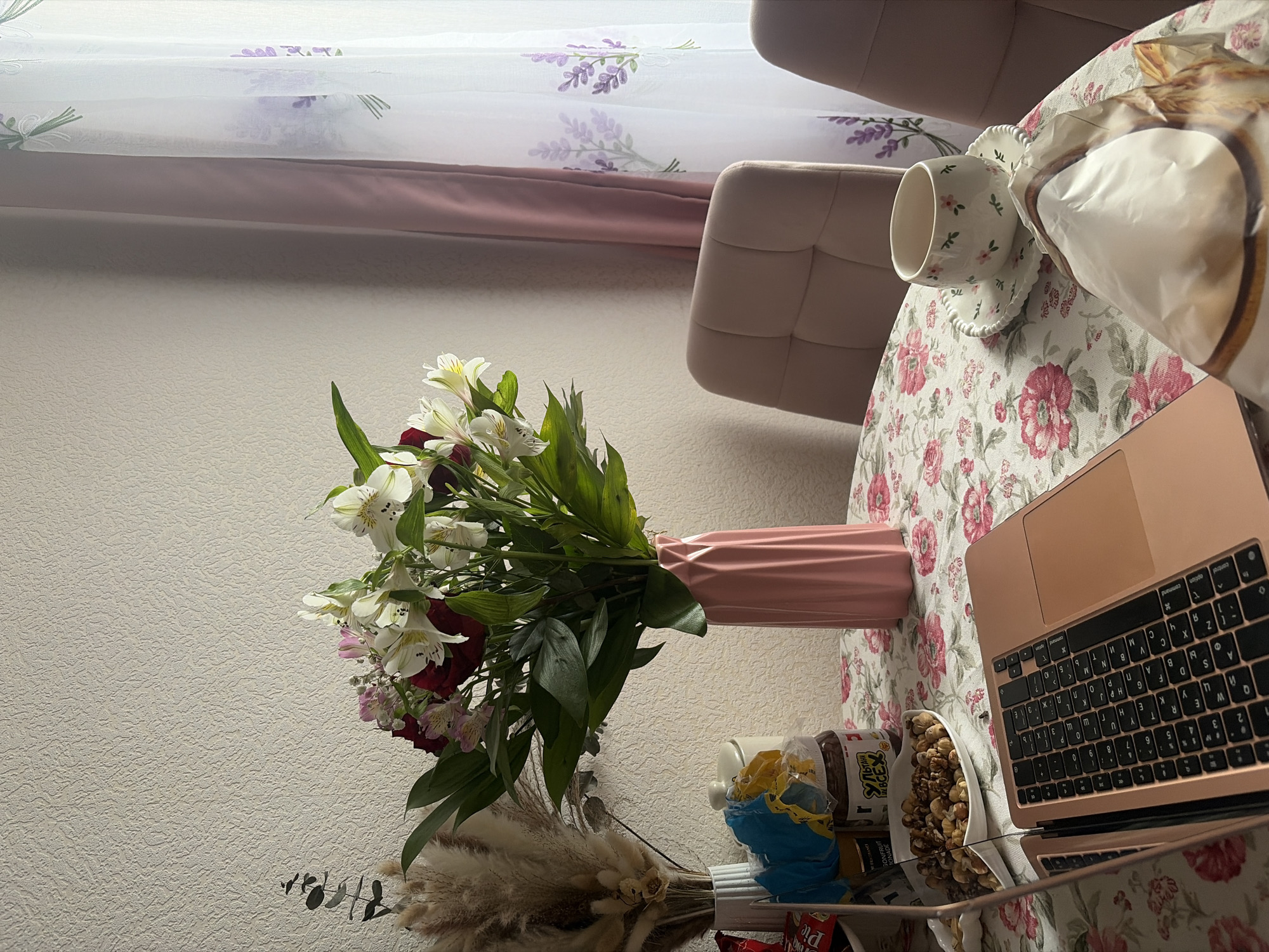 A still life featuring a bouquet of mixed flowers in a pink vase, set on a round table covered with a floral tablecloth. Adjacent to the vase is an open laptop. In close proximity to the laptop and vase are a cup and saucer, as well as containers holding food items like nuts. Behind the table is a chair with a beige upholstery and a window with a curtain featuring a lavender print. The background wall appears to be a pale color with a slightly textured surface. There are no people visible in the scene. This scene is not related to a specific location in Saint Petersburg, Russia.