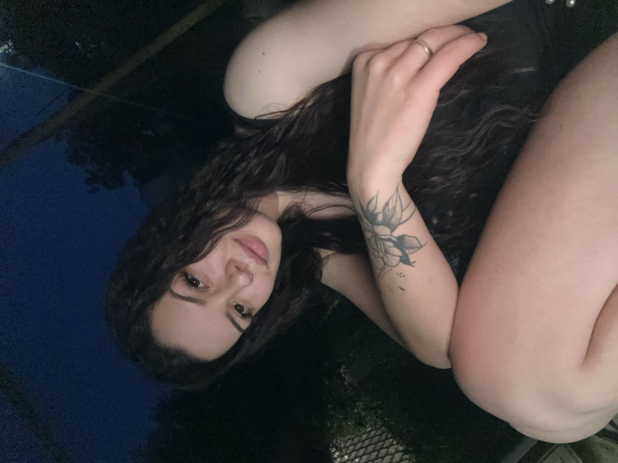 In Skala, Bulgaria, a young woman with long, dark, curly hair is pictured outdoors at night. She is wearing a black tank top. A tattoo of a floral design is visible on her left forearm. She has a ring on her right ring finger. The background is dark, with some trees and a fence visible. The lighting suggests artificial light is present, possibly from a nearby structure or a streetlamp. The woman is looking towards the camera with a relaxed expression.