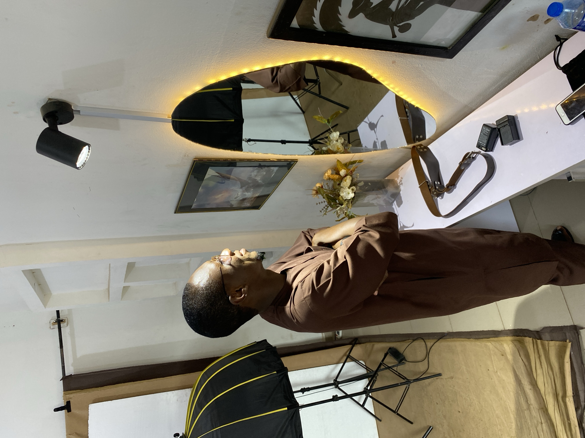 The image, rotated 90 degrees counter-clockwise, depicts an indoor setting, likely a photography studio or a room prepared for a photoshoot, in Bukuru, Nigeria. An adult male with dark skin, a shaved head, glasses, and a short beard stands with his arms crossed, looking upwards and to his left. He is wearing a long-sleeved, brown, collared garment, possibly traditional attire.
The room features white walls and a white ceiling with visible structural elements. A black spotlight is mounted on a track on the ceiling. Below it, an irregularly shaped mirror with a warm LED strip light around its edge is affixed to the wall, reflecting parts of the room's setup, including a large black and yellow photography umbrella on a stand. Two framed pictures adorn the wall; one with a landscape and another with a silhouette.
A white shelf runs along the wall, holding a small vase with artificial white and yellow flowers, two black battery packs (with small, unreadable text), a smartphone, a plastic water bottle with a blue cap, and a brown leather strap or belt. On the floor, a light-colored fabric or paper backdrop is spread out, with a white board and parts of another light stand visible.
The man appears calm and composed, suggesting he is posing or waiting for a photography session. Artificial lighting from the spotlight and the mirror's LEDs illuminates the scene.