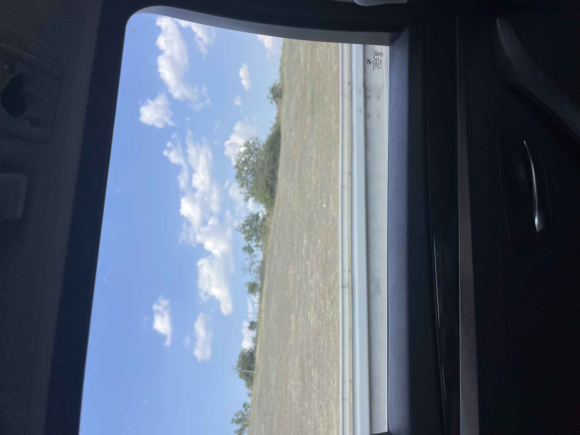 The media file is a photograph taken from inside a vehicle, looking out of the window. The scene outside is a rural landscape under a bright, sunny sky with scattered white clouds. The dominant colors are blue from the sky and a muted yellow-brown from a dry, grassy field. A line of trees, mostly green, runs along the edge of the field, suggesting a boundary or a distant treeline. A white barrier, possibly a guardrail or a fence, runs horizontally through the middle ground of the image.
The interior of the car is visible along the edges of the frame, primarily the dark-colored door panel. A metallic door handle is visible on the right side. The window itself shows some smudges or imperfections.
There is some text visible on the glass of the window, near the top right corner of the exterior view. It appears to be a series of symbols and letters, likely manufacturer or regulatory markings, including what looks like a BMW logo.
The lighting suggests it is daytime, likely midday, given the brightness of the sky and the clear visibility. The weather appears to be fair and sunny. The overall mood conveyed is one of travel and looking out at a peaceful, open landscape. The orientation of the image is vertical.