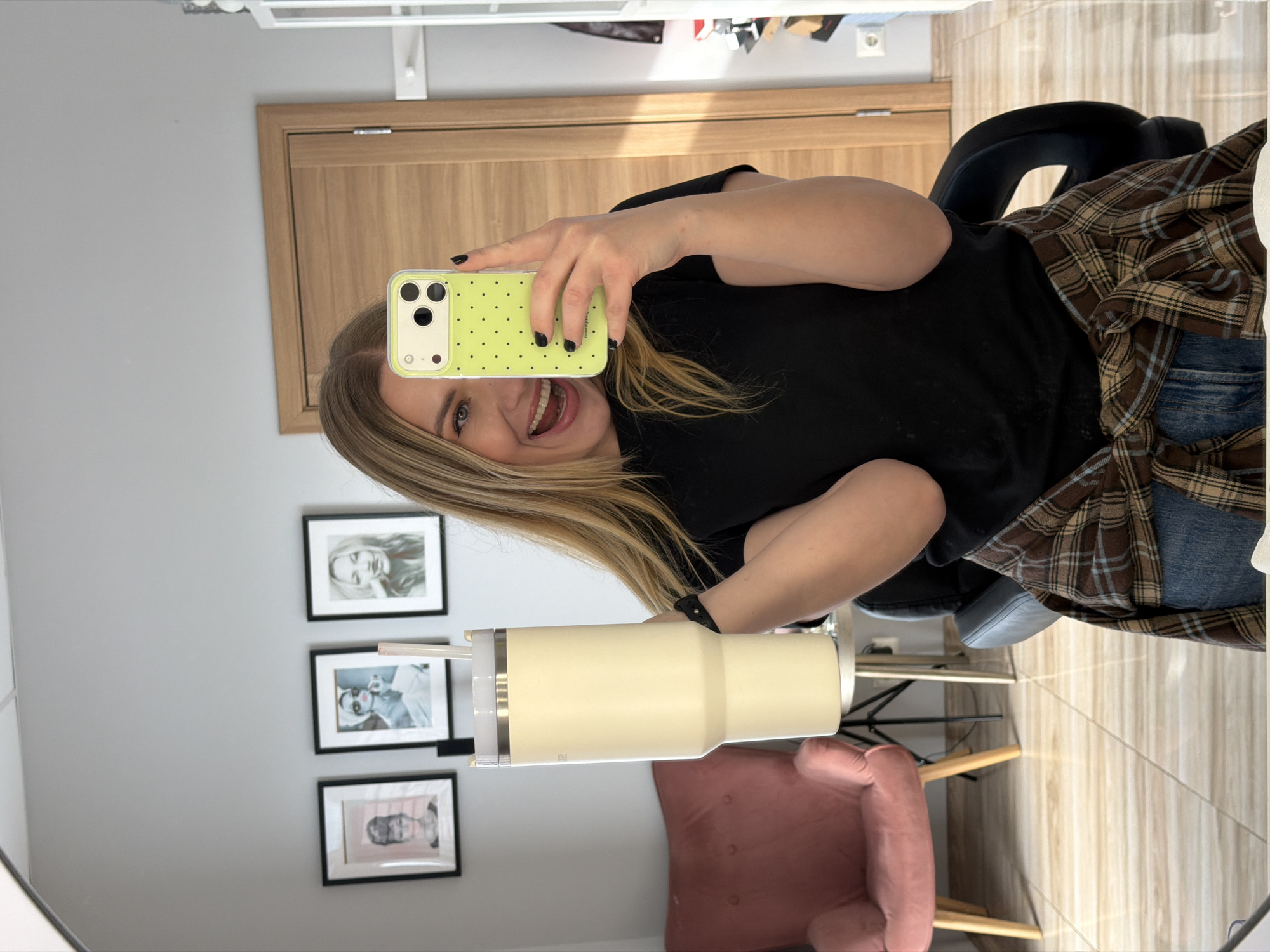 A selfie taken in a mirror shows a blonde woman smiling and holding a yellow phone with black polka dots. She is wearing a black t-shirt and a brown plaid shirt. She is holding a large, off-white tumbler with a straw. The mirror reflects a wall with three framed pictures and a pink chair. The background includes a light-colored wooden door and a glimpse of patterned floor tiles. The location appears to be an indoor space, potentially in Riga, Latvia, based on the provided contextual requirement.