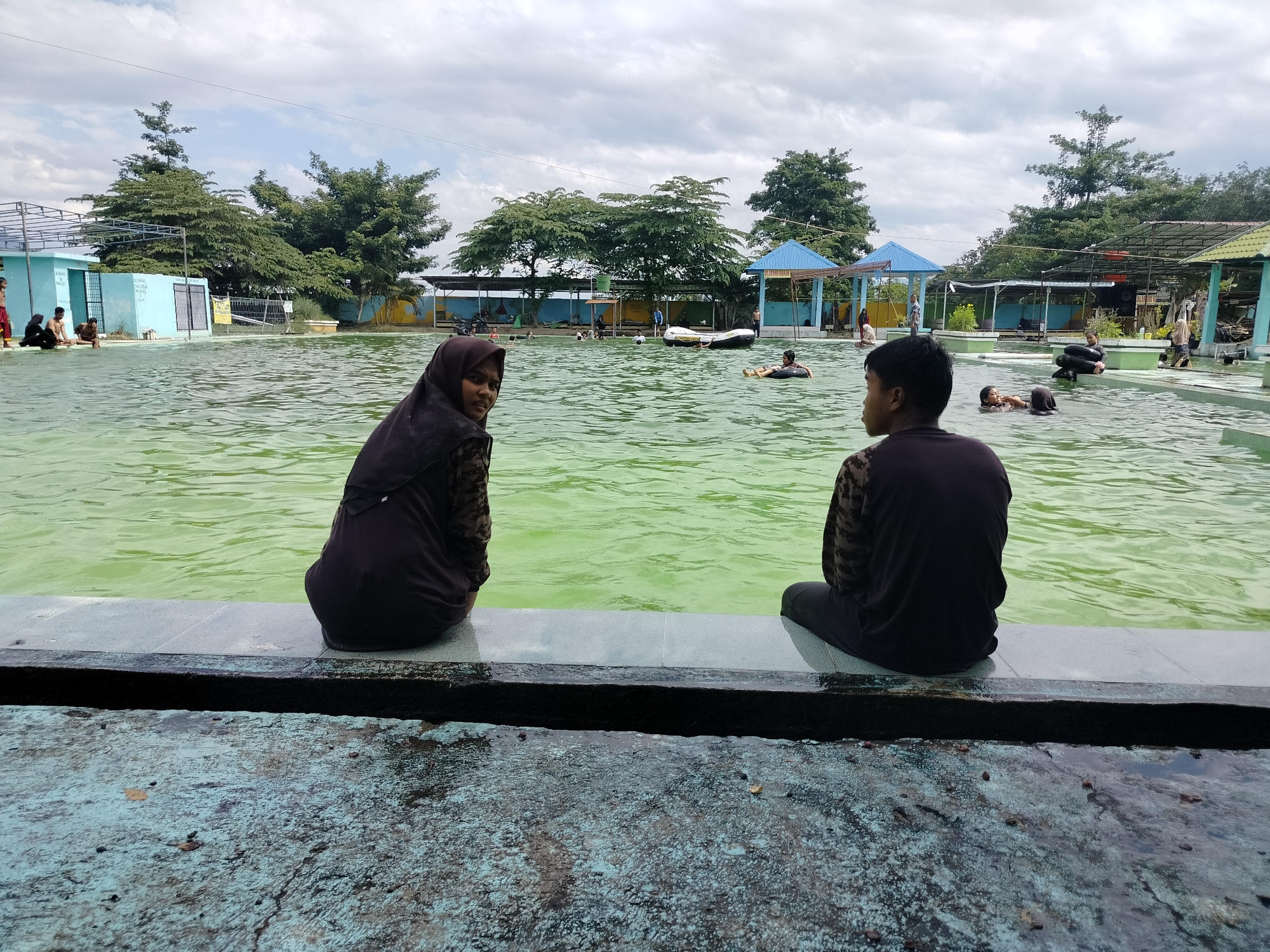 The image depicts a lively scene at a swimming pool complex, possibly a water park, in Tegarmiko, Indonesia. Two individuals are prominently seated on the edge of the pool, facing away from the camera and towards the water. The person on the left is wearing a dark hijab and patterned top, while the person on the right is wearing a dark, patterned shirt.
The swimming pool itself is filled with murky green water, and several other people are visible enjoying the water. Some are swimming, while others are floating on inflatable rings. In the background, there are various structures typical of a public pool, including changing rooms, shaded seating areas with blue and green roofs, and trees that provide a natural backdrop. The sky is overcast with clouds.
The foreground of the image shows a worn, textured concrete surface, suggesting it is part of the pool's deck area. The overall impression is of a recreational facility bustling with activity on a cloudy day.