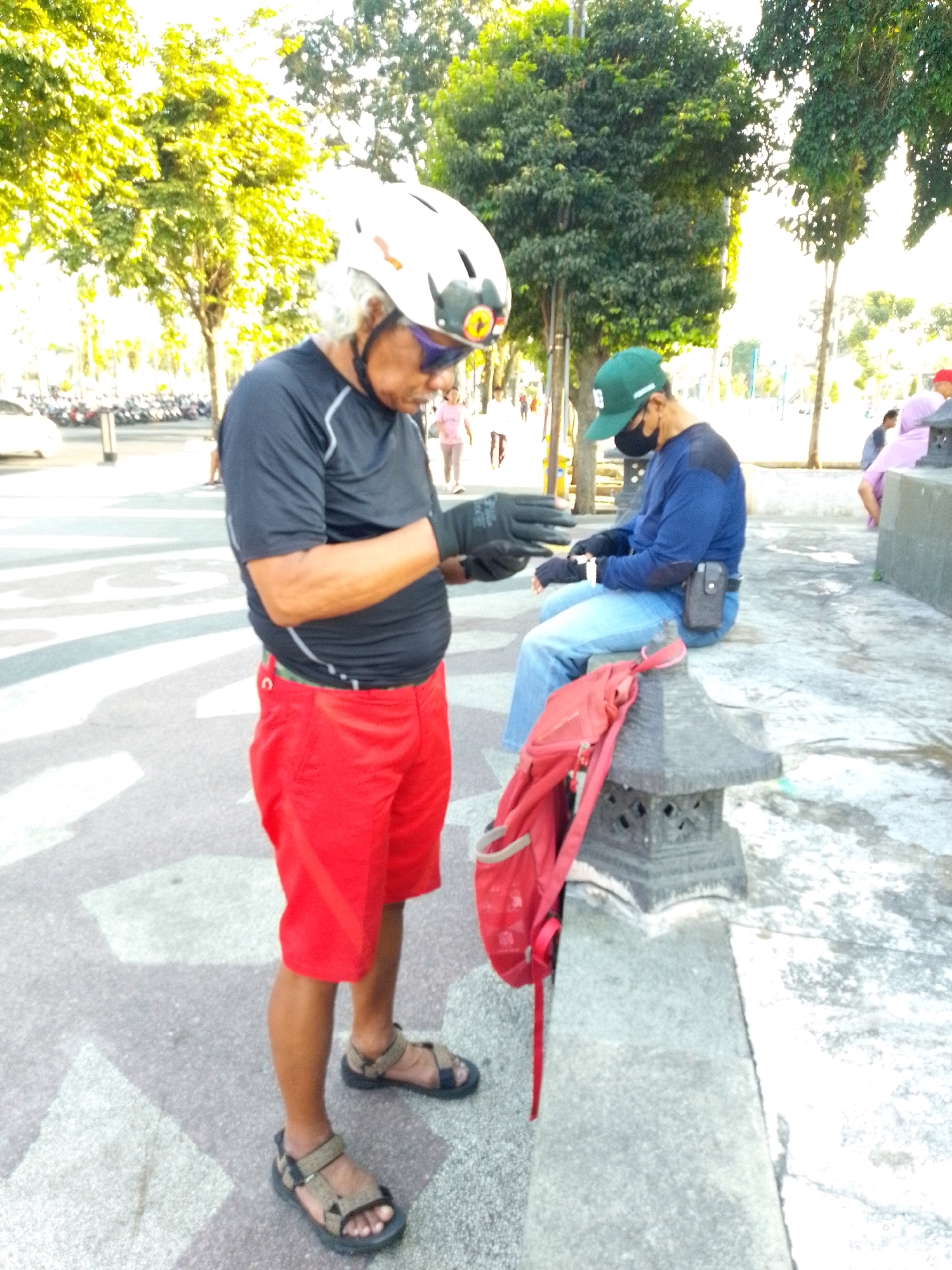 In Jember, Indonesia, an older man with white hair stands on a street, wearing a black long-sleeved shirt, red shorts, black gloves, a white helmet, and sunglasses. He appears to be interacting with his hands. A younger person in a blue long-sleeved shirt, blue jeans, and a green cap sits on a concrete ledge, looking down. A red backpack rests on a decorative stone structure near the sitting person. The environment features trees, parked motorcycles, and street markings.