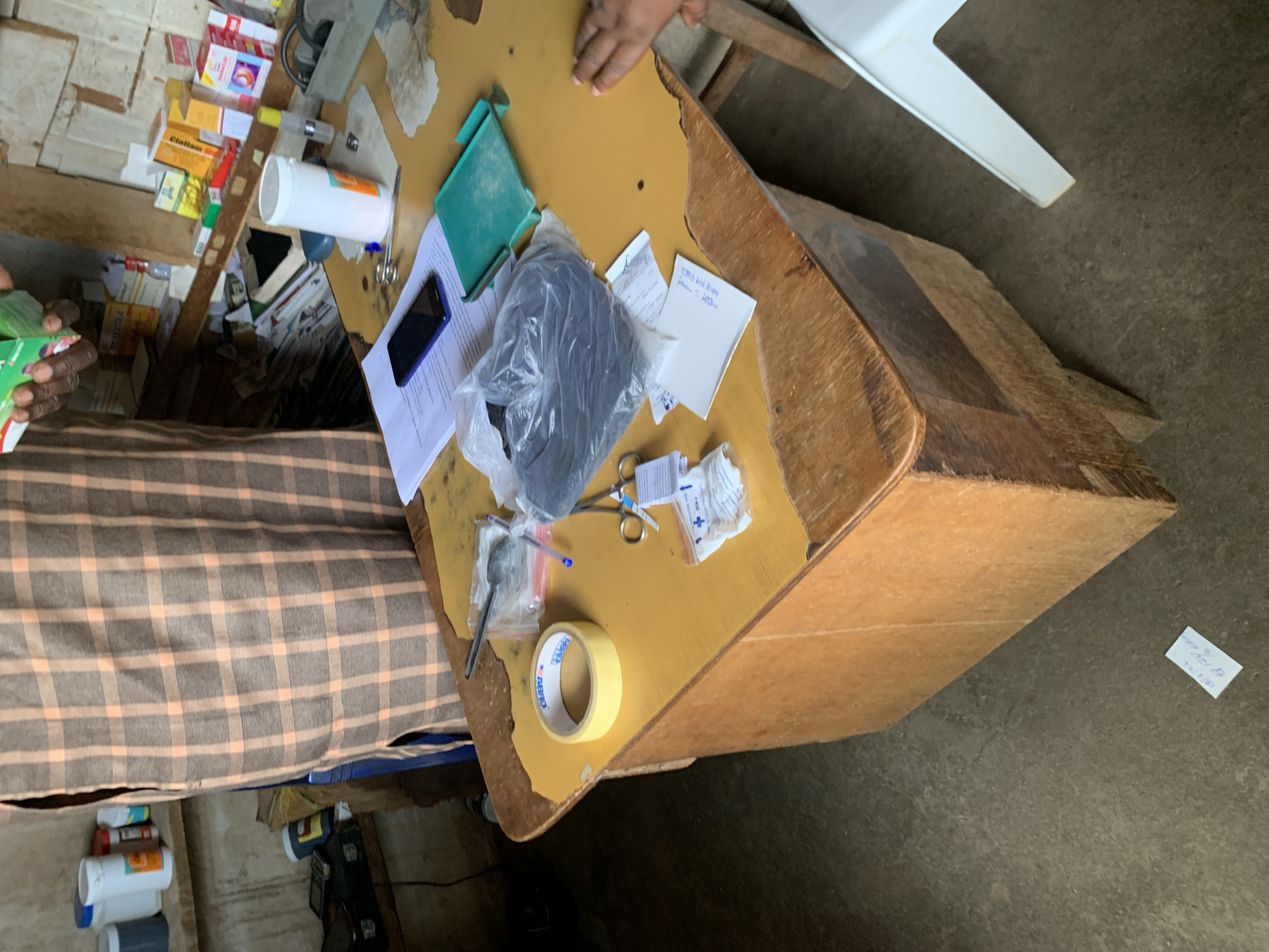 The image shows a close-up view of a wooden desk in what appears to be a small shop or clinic. The desk is cluttered with various items, including a roll of yellow tape, scissors, small plastic bags containing what looks like medical supplies, a smartphone, a white cylindrical container, and a green plastic tray. In the background, shelves are stocked with boxes of medications. A person wearing a brown and orange checkered garment stands behind the desk, holding a green box of medication. The floor is concrete, and a white plastic chair is partially visible on the right side of the image. The scene is likely in the city of Jos, Nigeria, given the context of the provided additional information which I am not supposed to use.