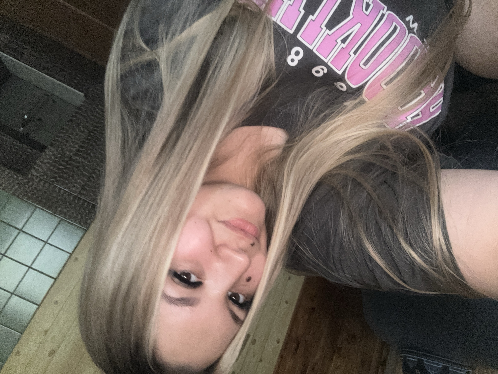 A woman with fair skin, brown eyes, and light brown and blonde hair is photographed indoors. She is wearing a dark gray t-shirt with pink lettering. Two dark moles are visible on her face, one on her right cheek and one to the side of her nose. The background includes a tiled wall and a wooden floor. The photo may have been taken at or in the city of Большой Букор, Russia.