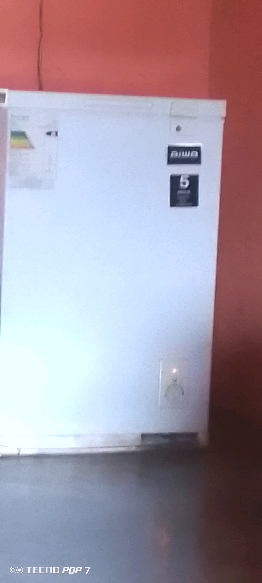 The image shows the side of a white chest freezer with an "AIWA" logo and a rating of "5" visible on its surface. There is an energy efficiency sticker on the left side of the freezer and a power outlet with a red indicator light on the right side. The freezer is placed against a salmon-colored wall. The lower portion of the image is dark, suggesting it is a low-light environment. A watermark "TECNO POP 7" is visible in the bottom left corner, indicating the device used to capture the image. The scene appears to be an indoor domestic setting. There is no discernible information in the image that would allow for the specific identification of its location within Altagracia, Venezuela.