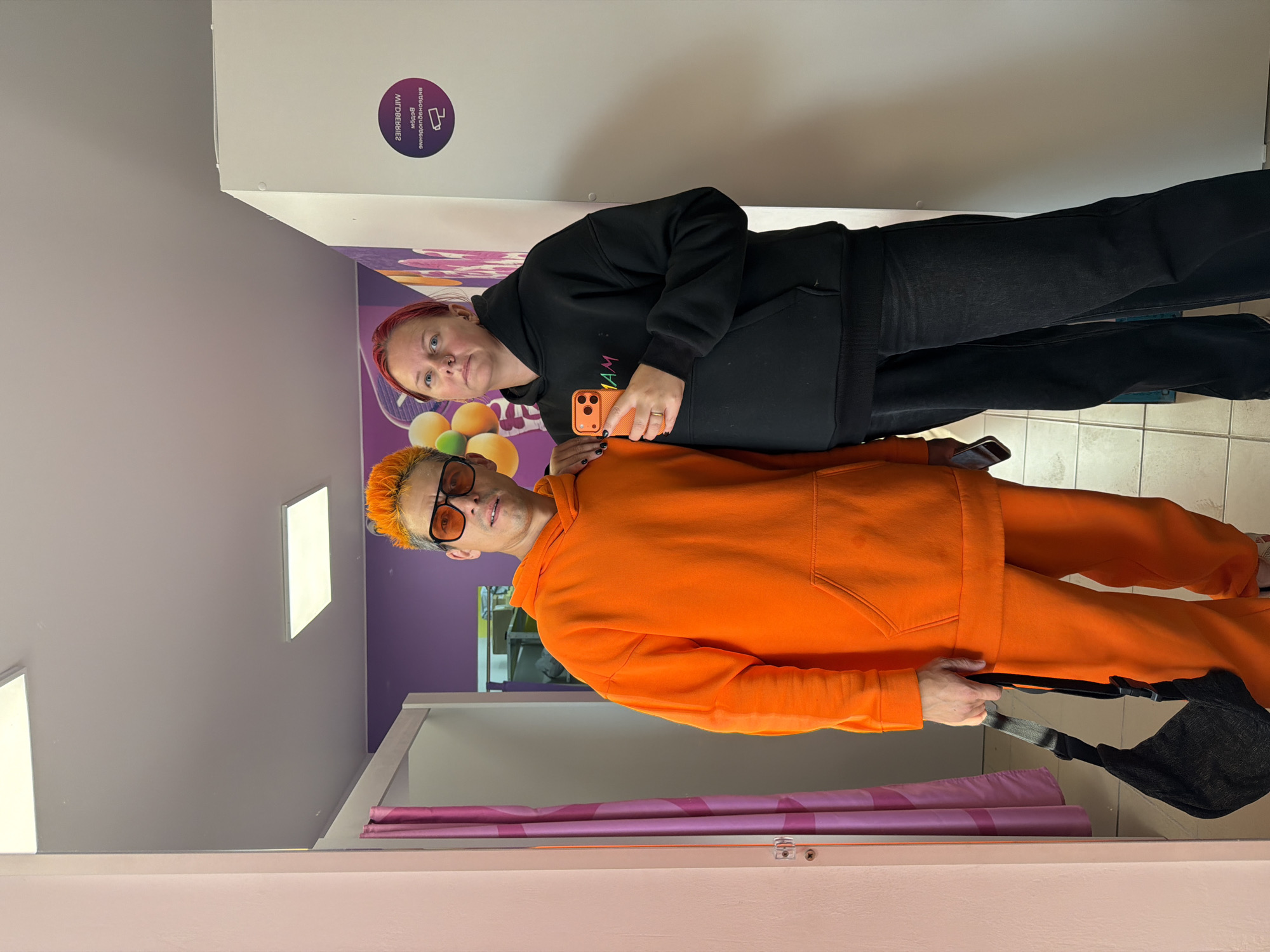 The image shows a man and a woman standing in a fitting room. The man has bright orange hair and is wearing an orange hoodie and pants. He is also wearing orange sunglasses and carrying a black bag. The woman has pink hair and is wearing a black hoodie and black jeans. She is holding a smartphone. The fitting room appears to be in a retail store, possibly a clothing store, as there are fitting room stalls visible. There is a circular sticker on the wall with text in Russian, which appears to be a store logo or information. The floor is tiled. The overall impression is a candid moment captured in a commercial setting, likely in Chaikovsky, Russia.
