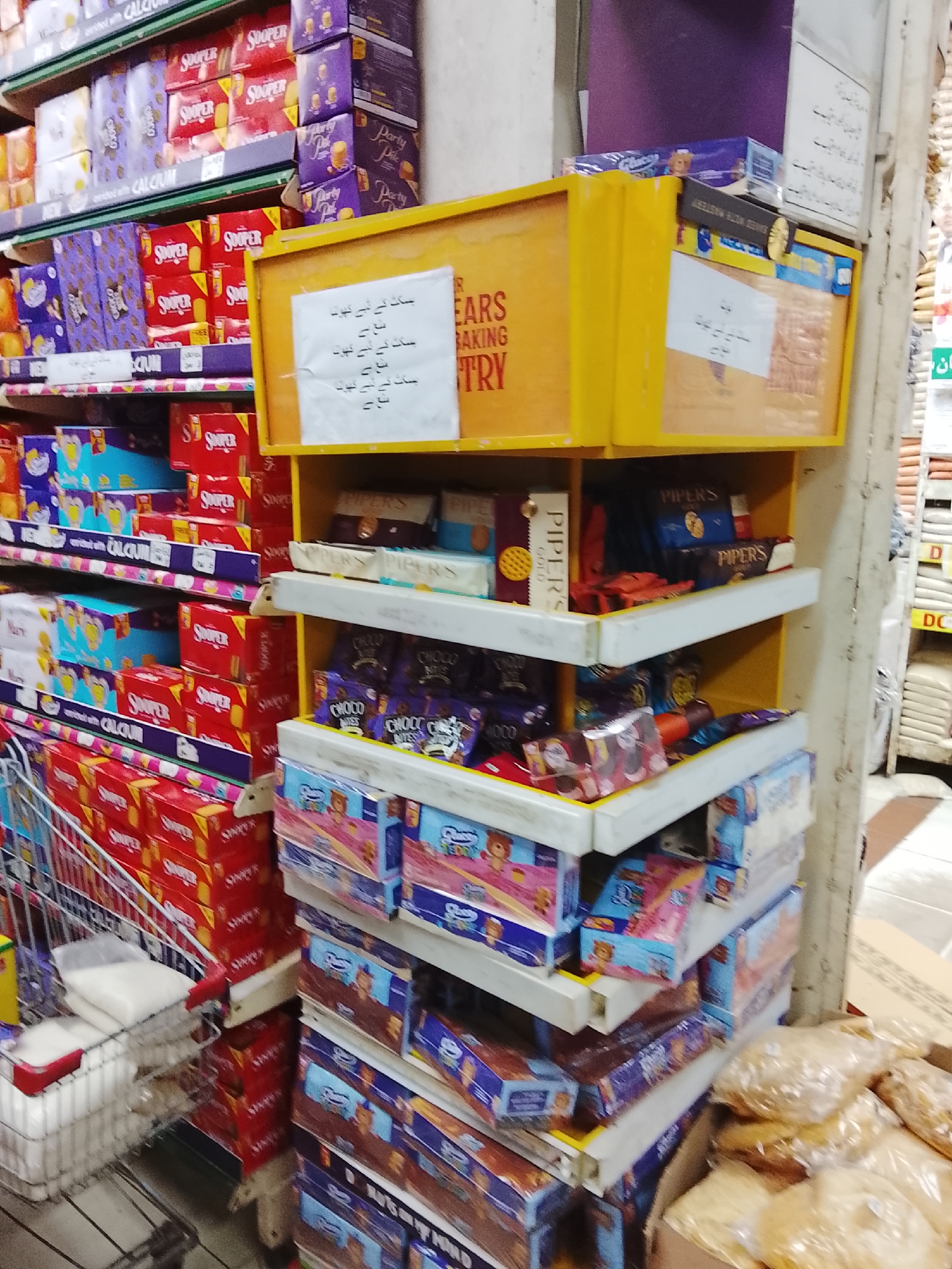 The image shows a grocery store shelf stocked with various biscuit and snack products. Dominant brands include "Sooper" and "Pipers." A yellow display unit stands out, showcasing more "Pipers" products. The shelves are densely packed, and a shopping cart with items is visible to the left. The environment appears to be a typical grocery store setting.