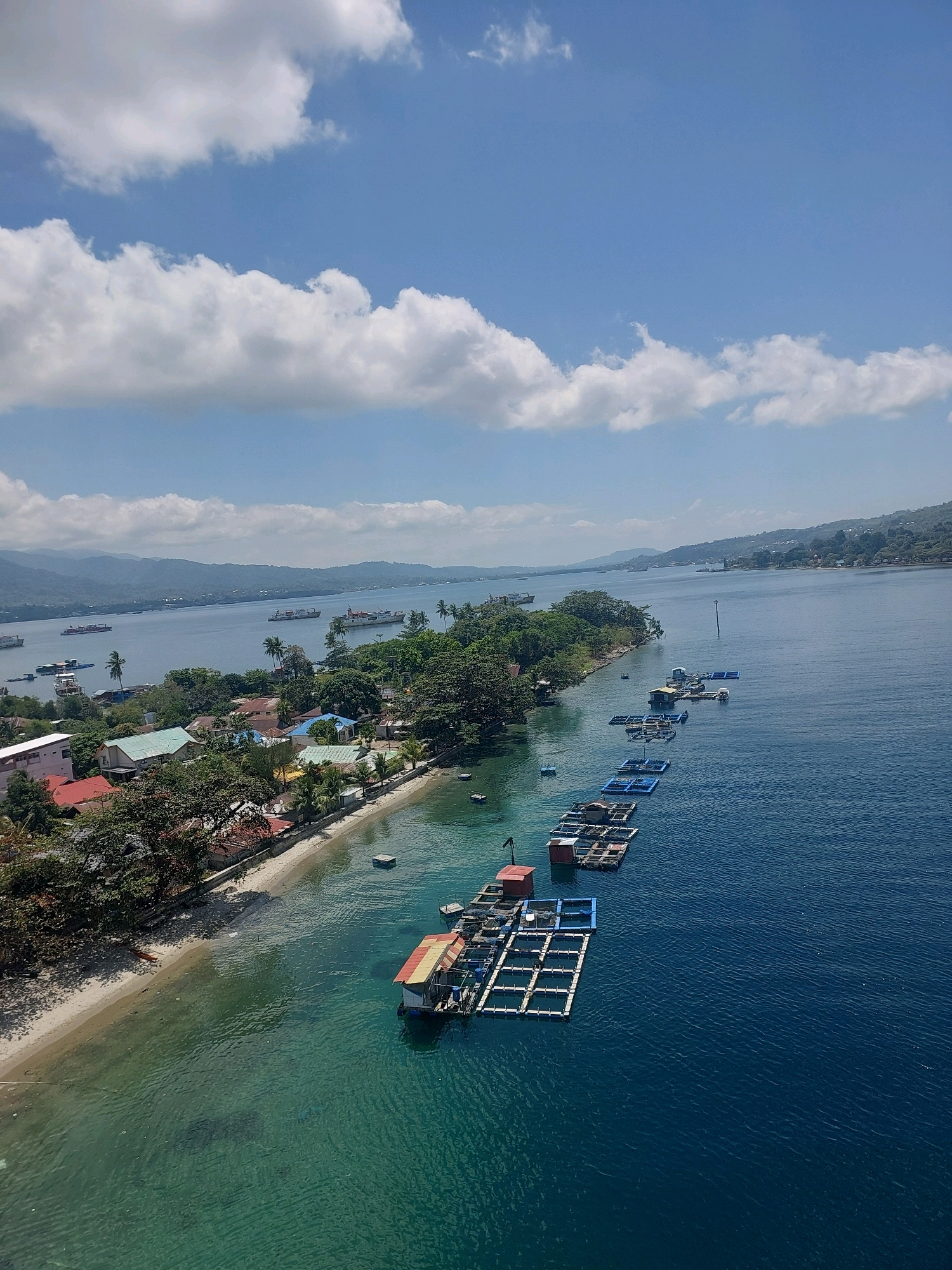 The image shows a coastal scene in Ambon, Indonesia, with the foreground featuring a clear turquoise body of water. Along the water's edge is a sandy beach backed by a dense area of green trees and various buildings with multi-colored roofs, suggesting a residential or small commercial area. Further out into the deeper blue water, a series of floating fish farms are visible, arranged in a long, linear pattern. These farms consist of numerous rectangular cages, some with small structures on top. In the background, the expansive body of water extends to a distant shoreline with hills and mountains, under a bright blue sky dotted with fluffy white clouds. Several large ships are anchored or moving in the distance, indicating maritime activity. The overall scene depicts a tranquil coastal environment with both natural beauty and human infrastructure.